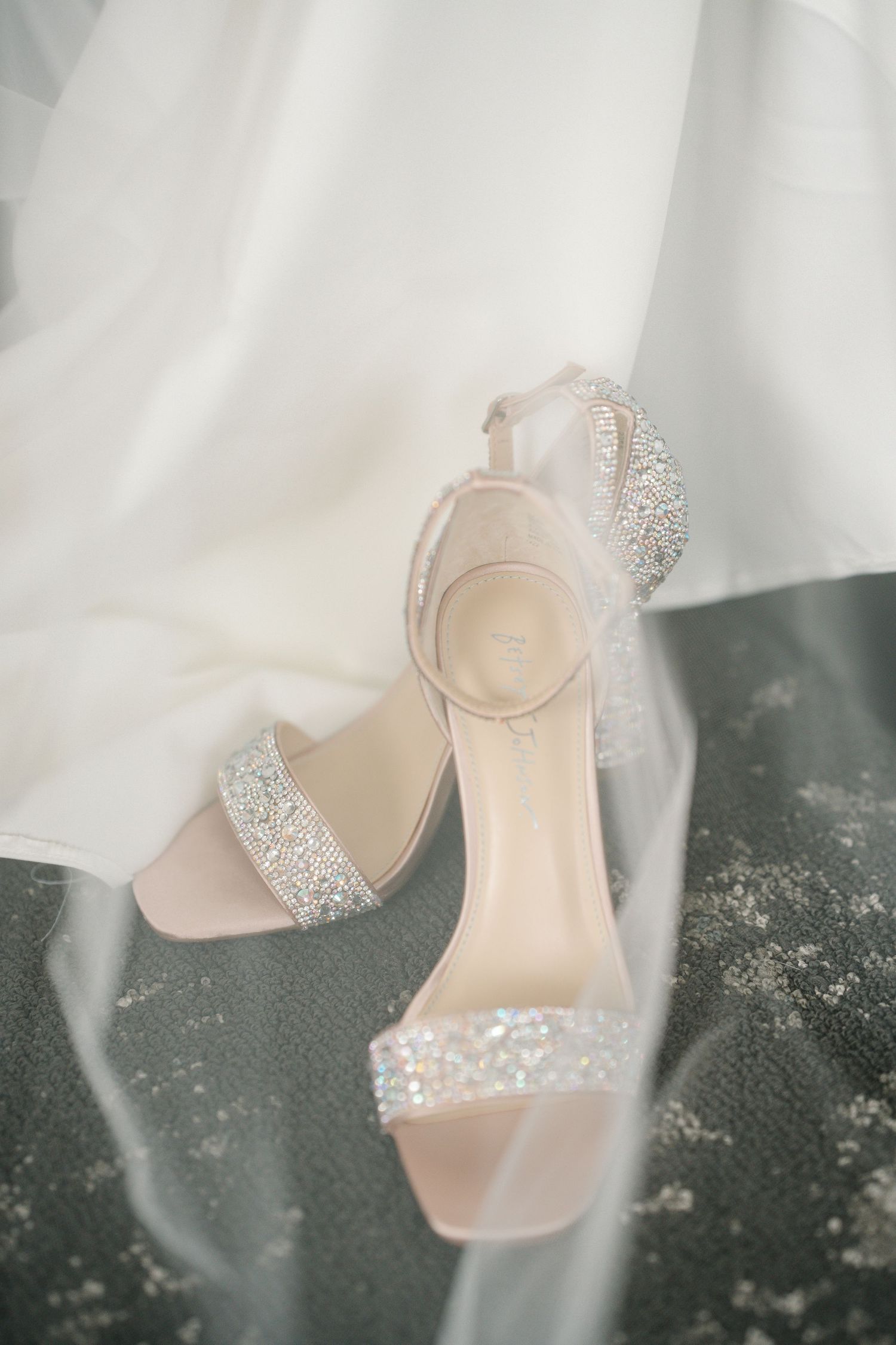 Elegant blush pink bridal shoes with sparkling crystal embellishments against a soft white fabric backdrop.