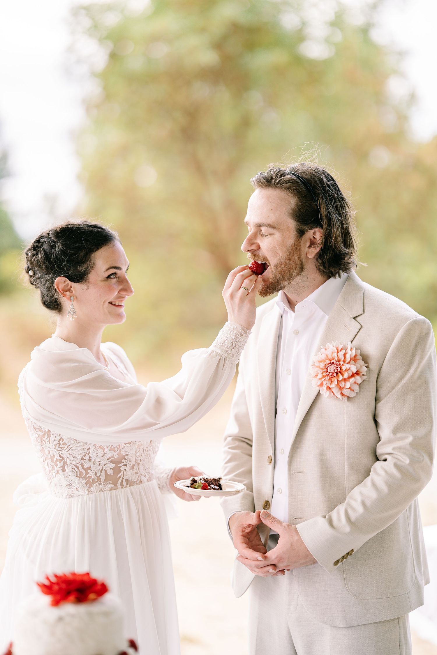 A romantic wedding couple in white attire share joyful candid moments in a sun-dappled outdoor setting.