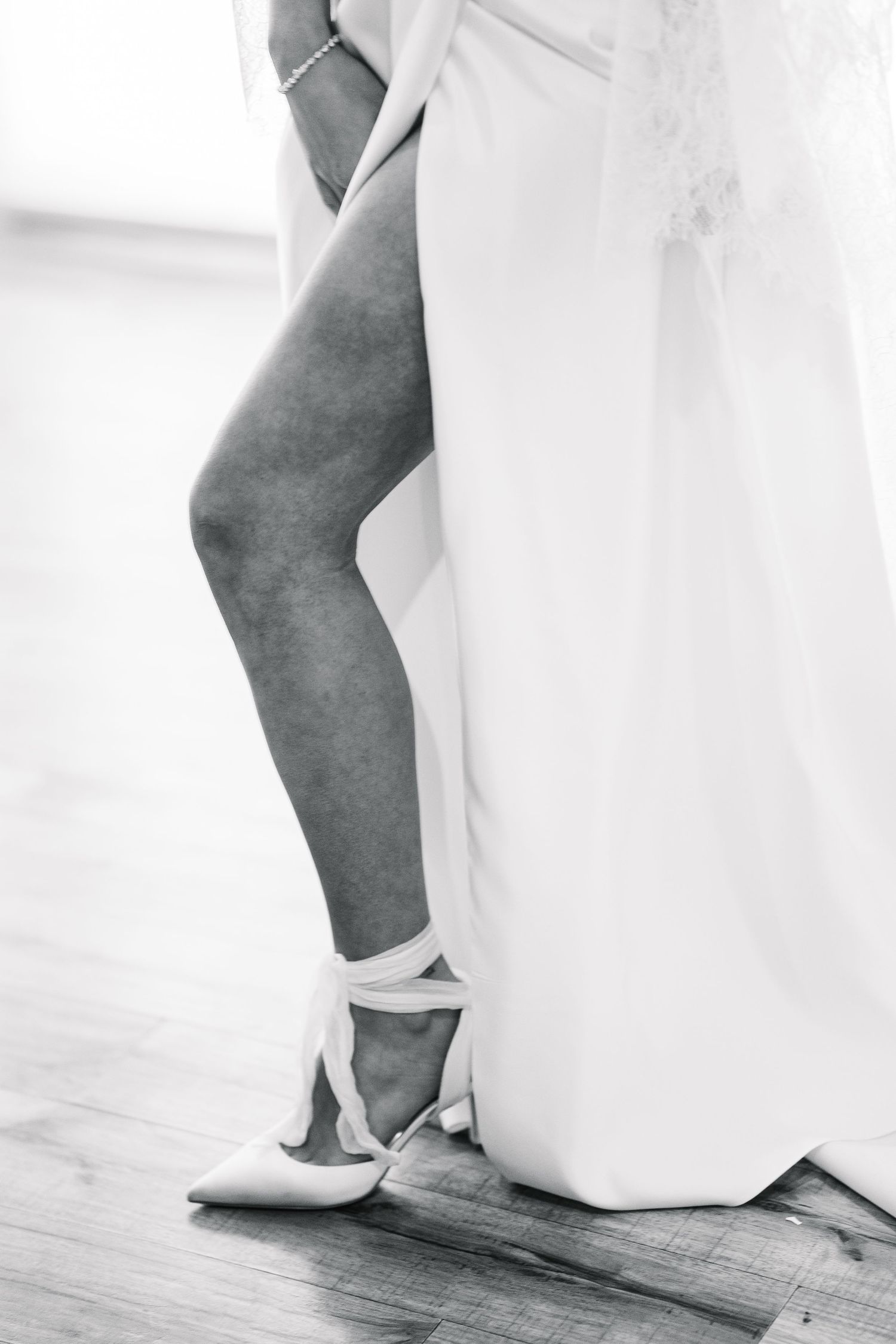 A bride's feet in elegant white heels stand on a wooden floor next to the flowing hem of her wedding gown.