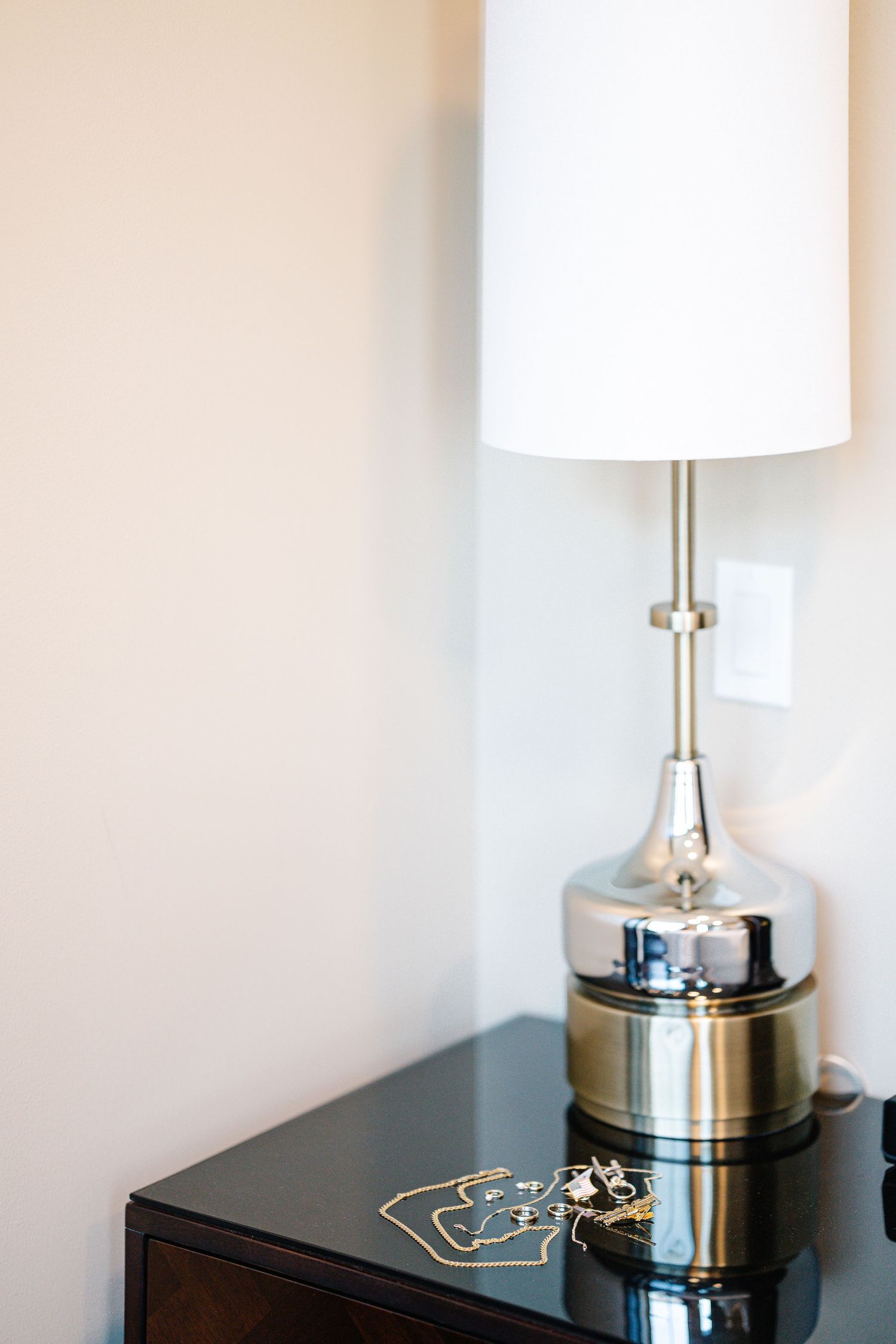 Modern table lamp with white shade and metallic base sits on dark furniture against neutral wall.
