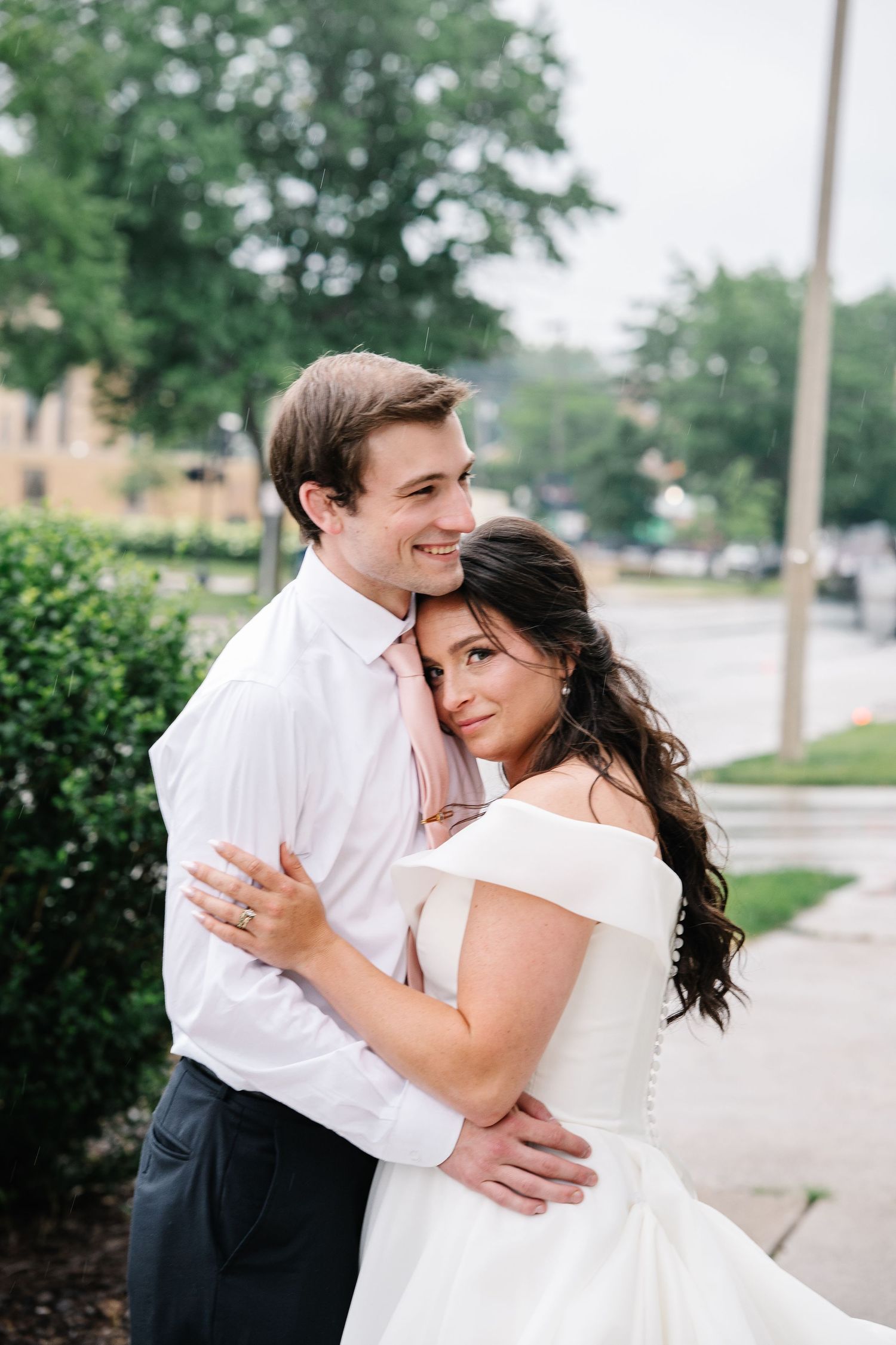 A couple embraces outdoors on their wedding day, with the bride in a white off-shoulder dress and groom in white shirt.
