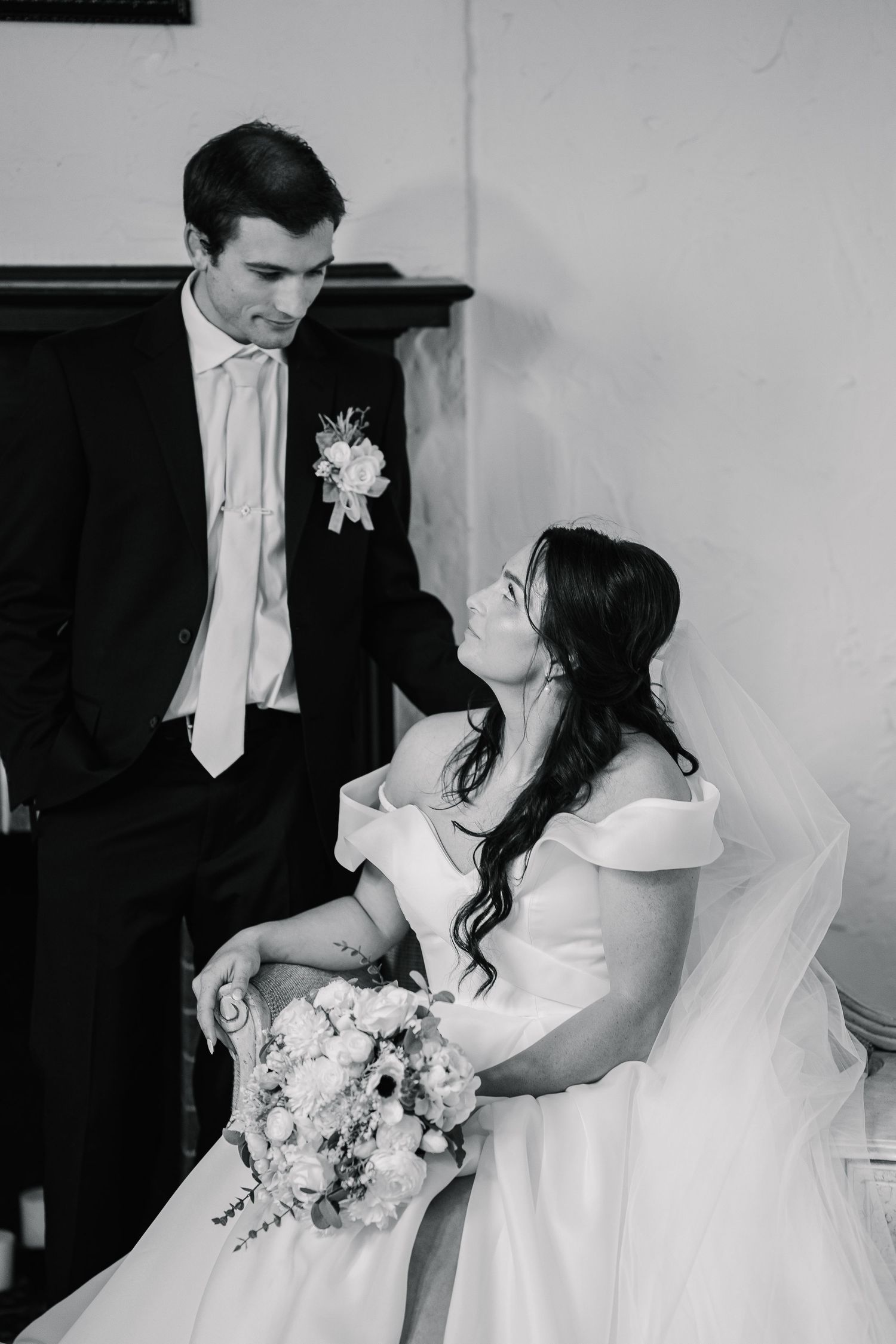 Black and white wedding photo of bride in off-shoulder gown looking up at groom in tuxedo with boutonniere.