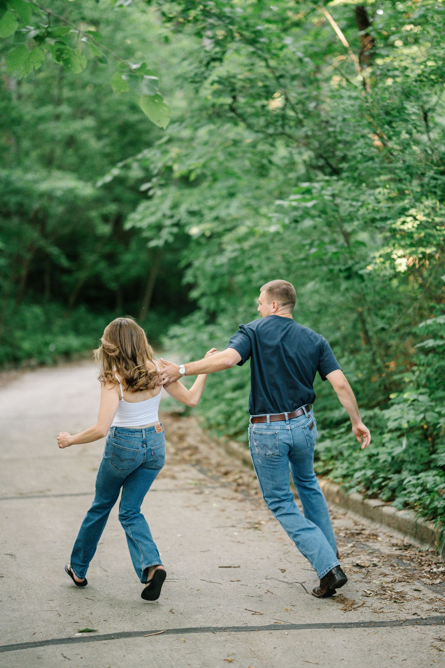 Couple holding hands running down a tree-lined path in jeans and casual tops during an outdoor photo session.