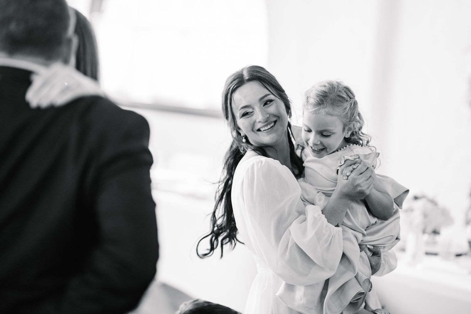 Black and white candid photo of a woman in a white dress holding a small child during a celebration.