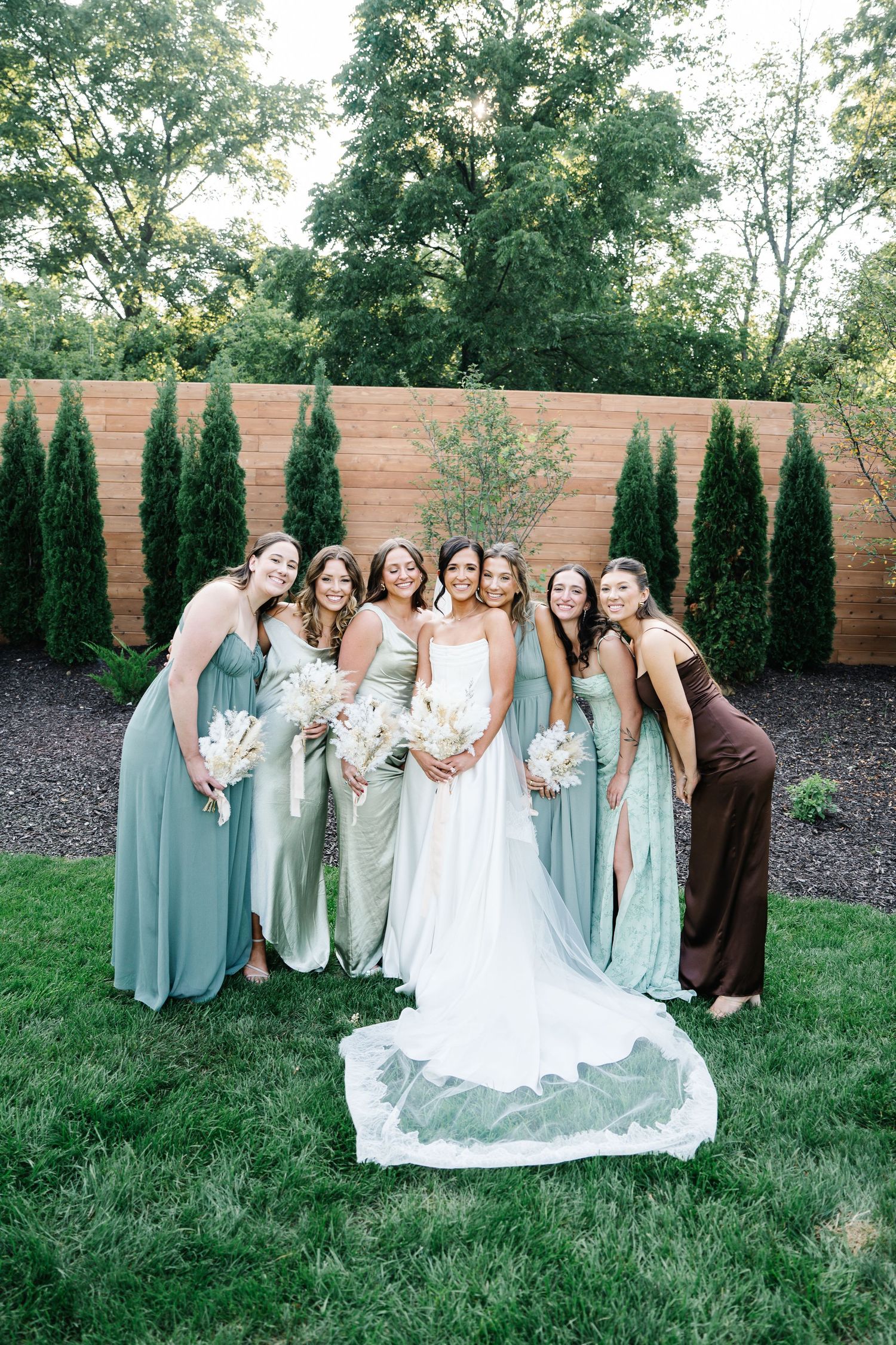 Wedding party in sage green dresses poses together on grassy lawn with wooden fence and evergreen trees in background.