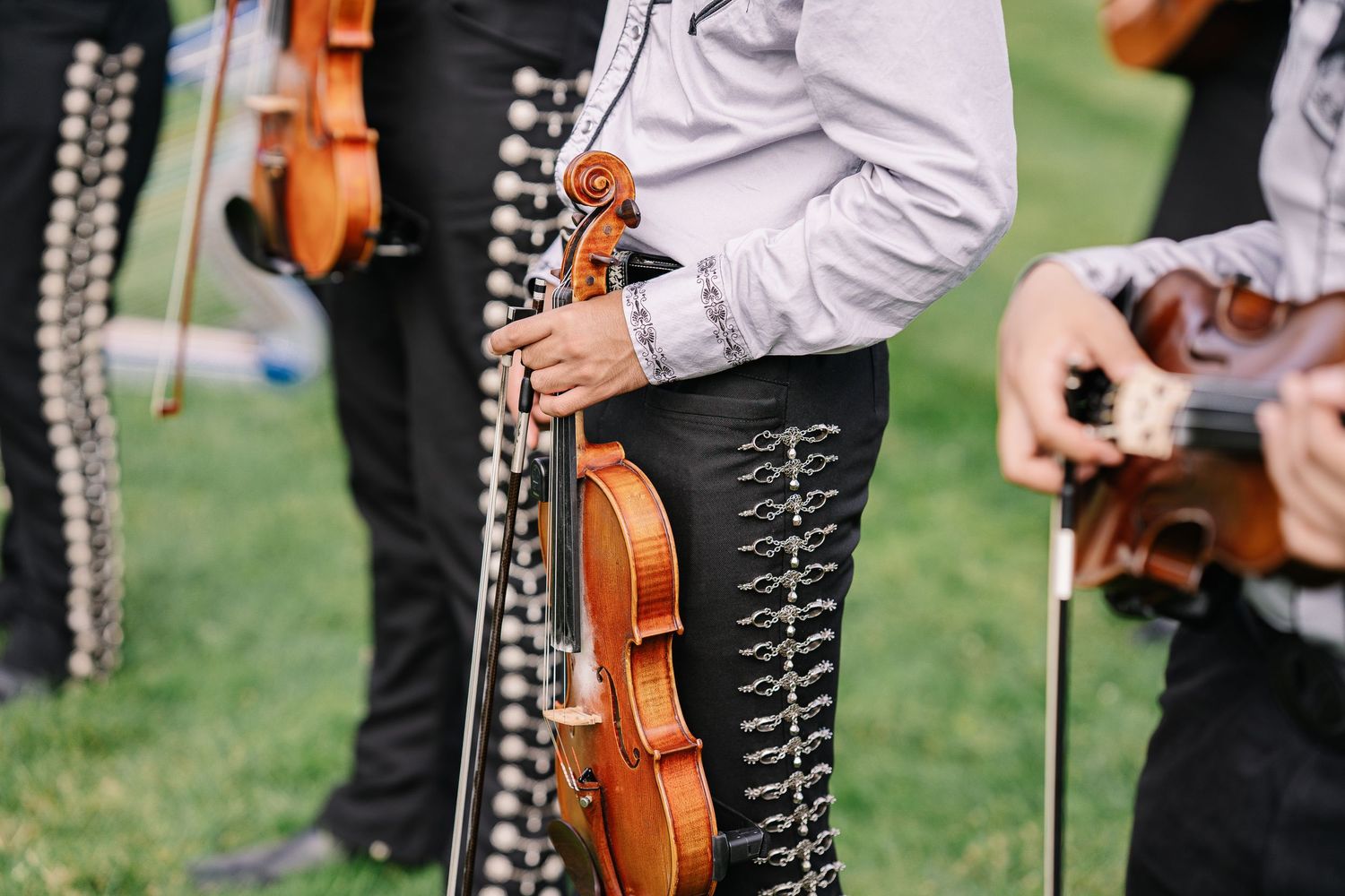 Musicians in traditional mariachi attire hold violins during an outdoor performance on grass.