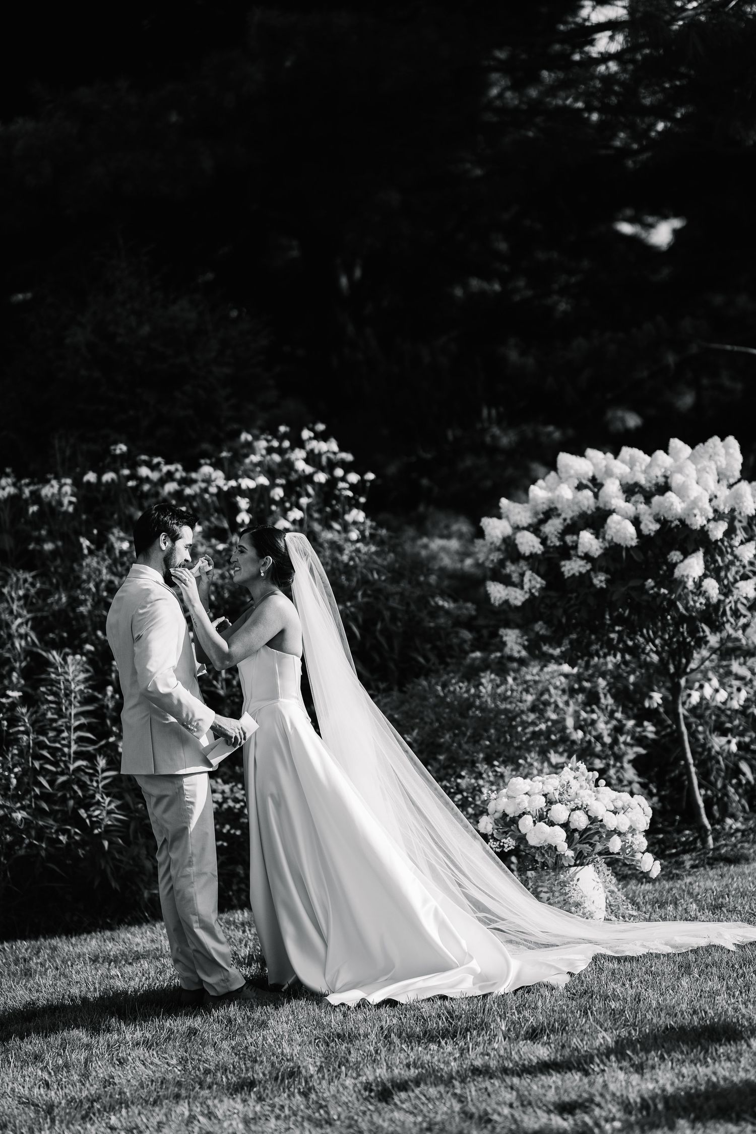 Black and white photo of wedding dresses arranged on grass with white flowers in the garden background.