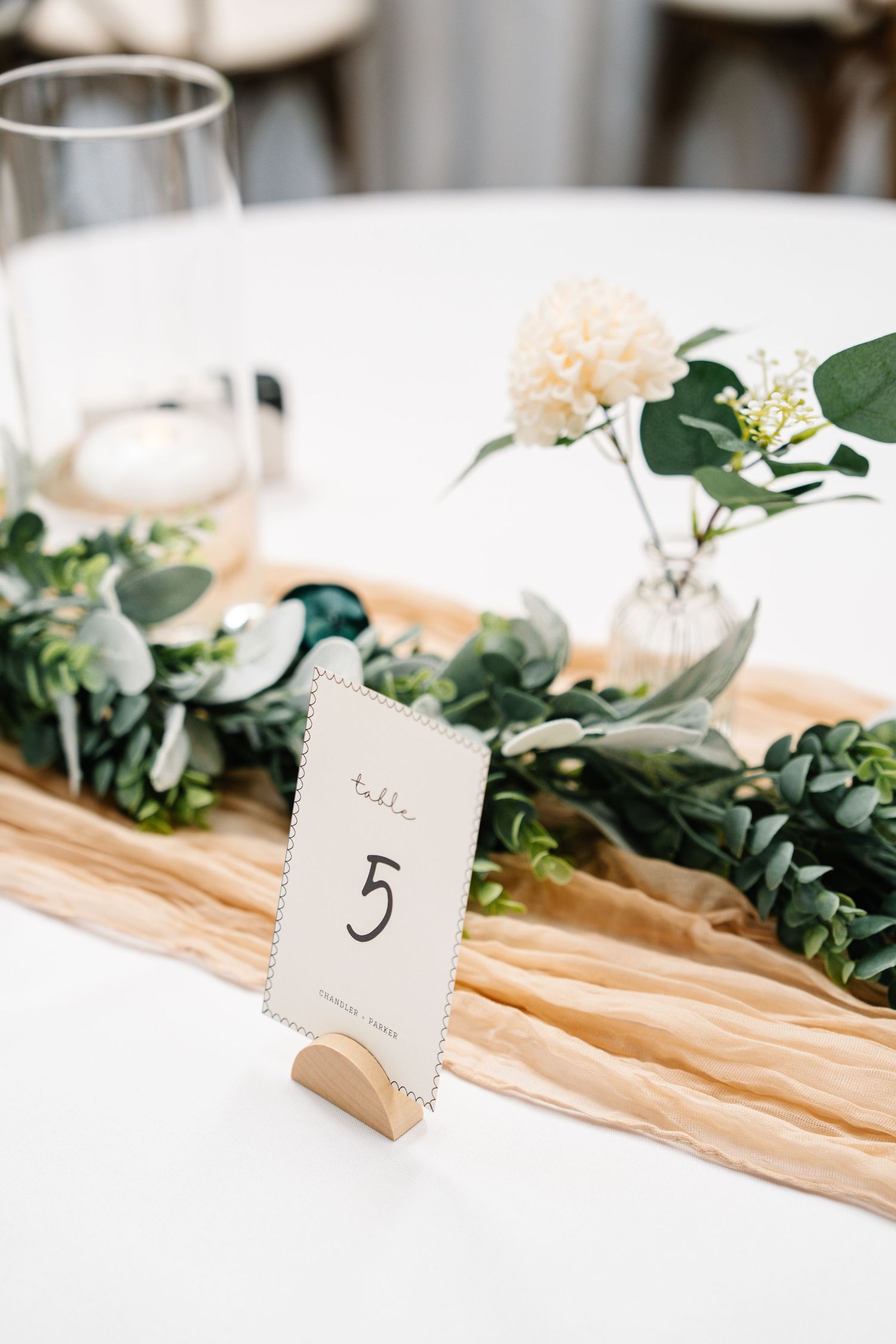 Minimalist table decor features eucalyptus garland and numbered table cards on light fabric runners.