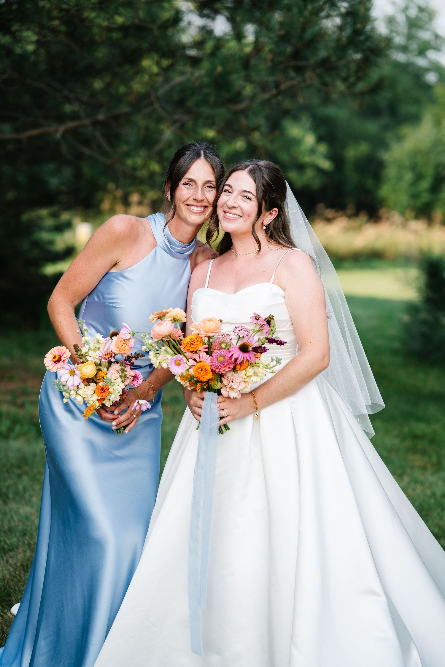 A bride in a white gown and bridesmaid in a light blue dress pose together with vibrant floral bouquets outdoors.