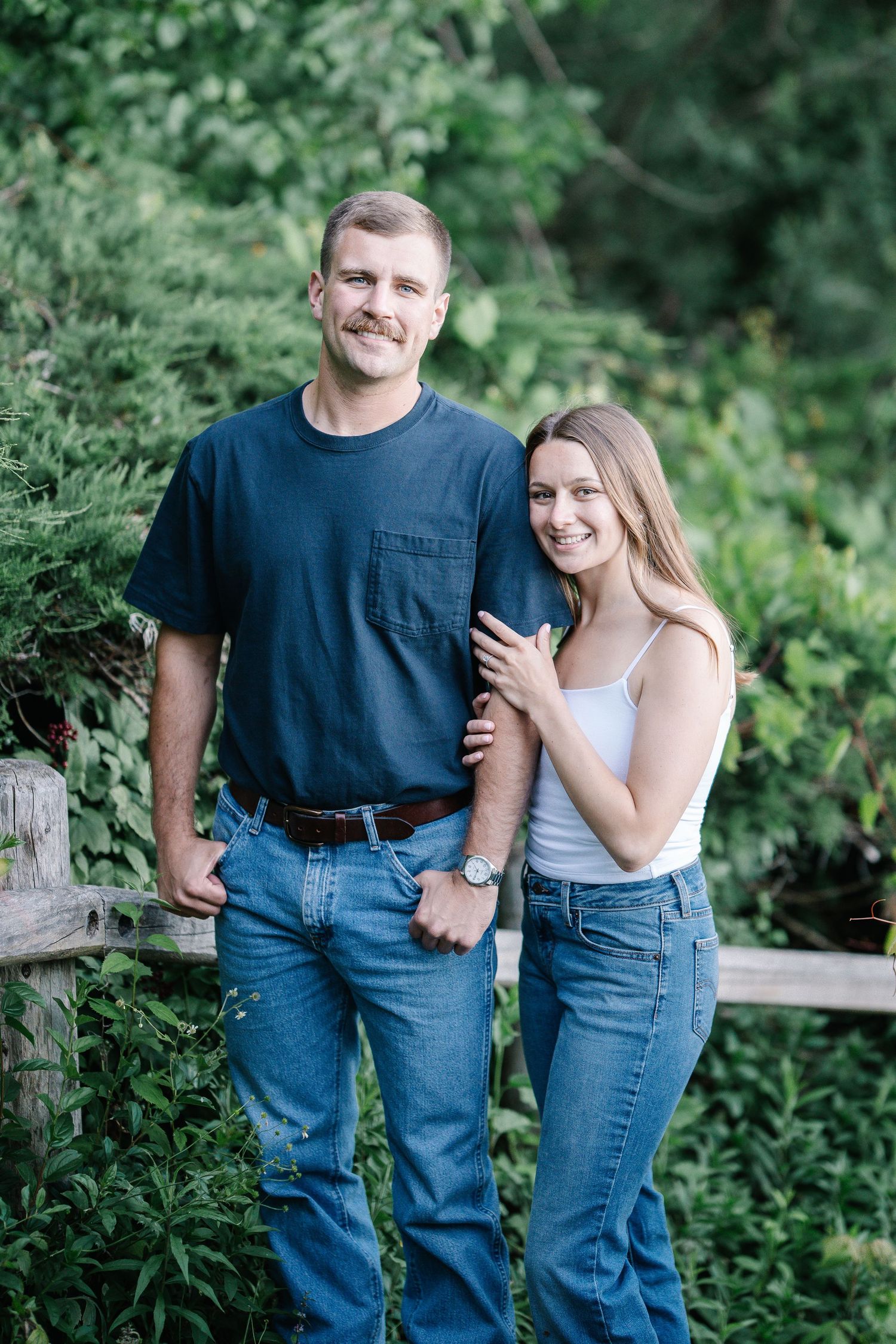 A smiling couple poses outdoors near a wooden fence surrounded by lush green foliage, both wearing jeans.