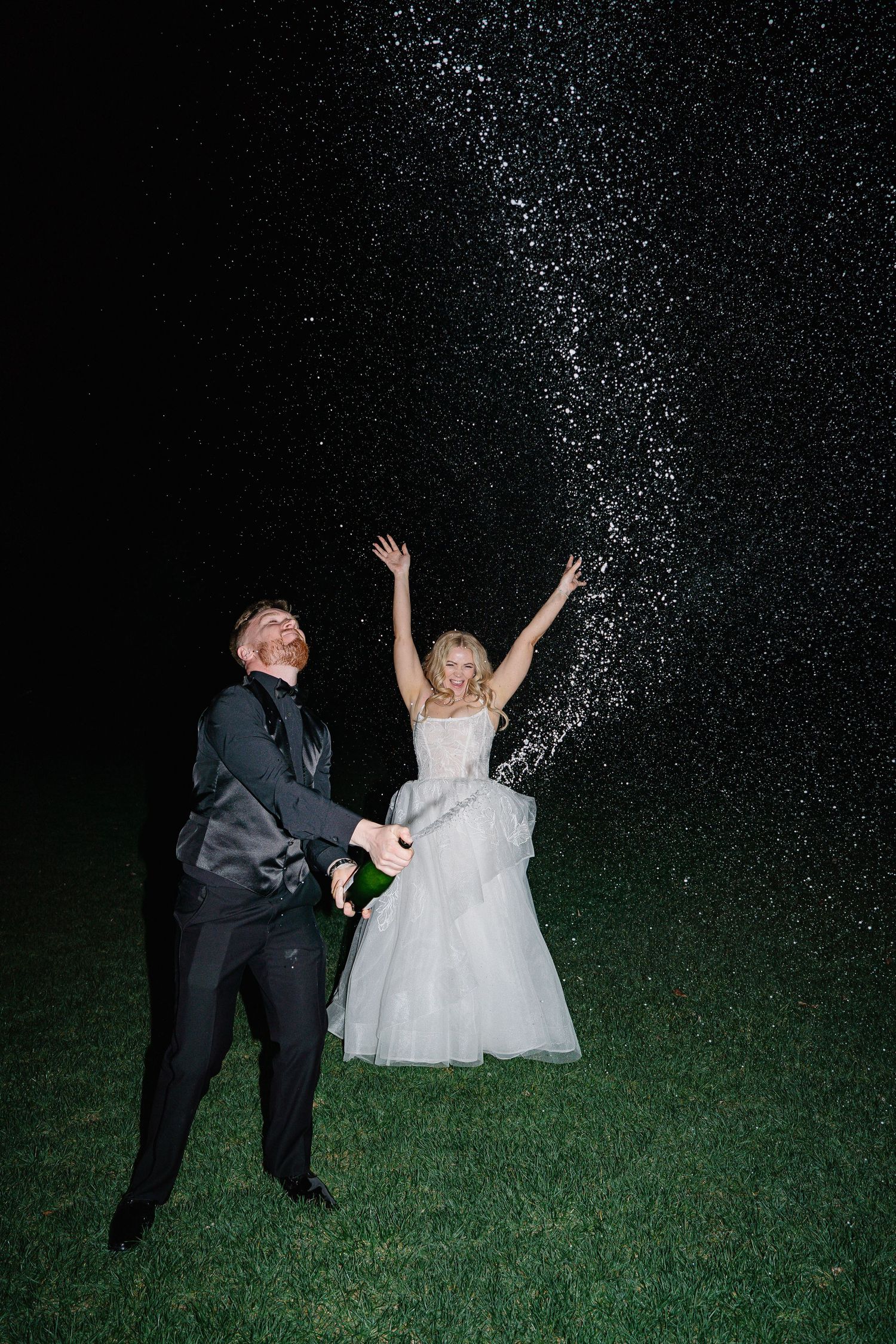 A couple in formal attire runs through sparkling nighttime sprinklers on grass during a celebratory moment.