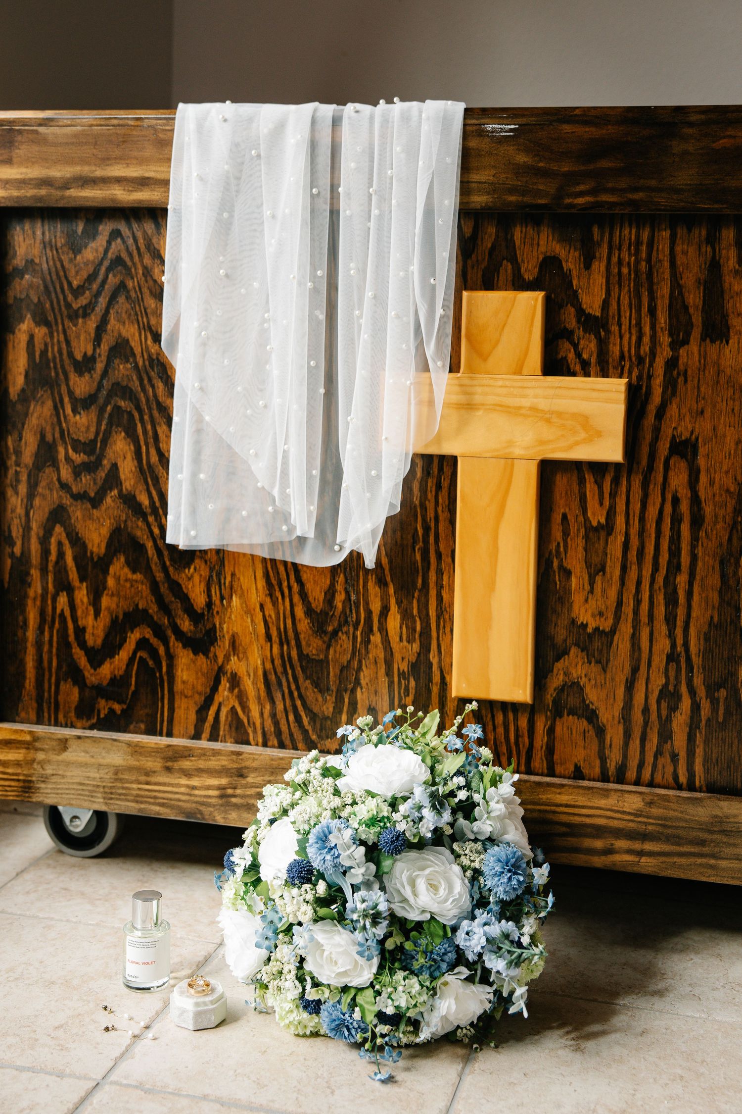 A wooden cross with white and blue floral arrangements sits against a rustic wooden wall with sheer white curtains.