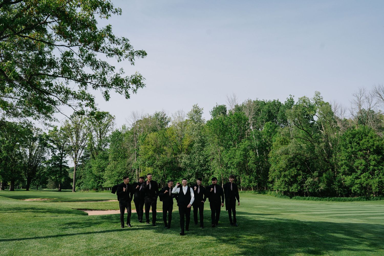 A group in formal black attire stands together on a lush green lawn surrounded by trees.