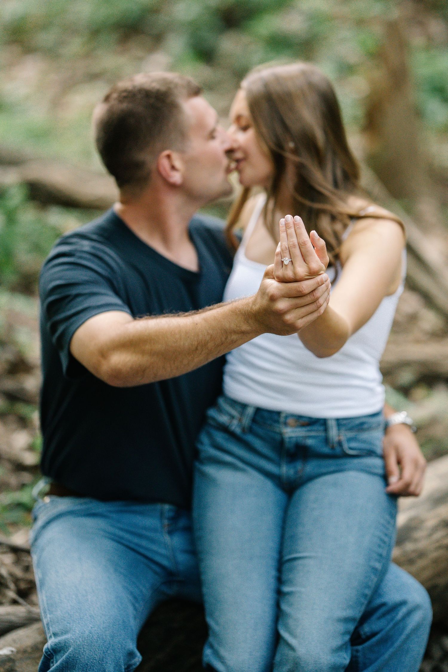 Couple sharing a kiss outdoors while showing off an engagement ring in a lush forest setting.