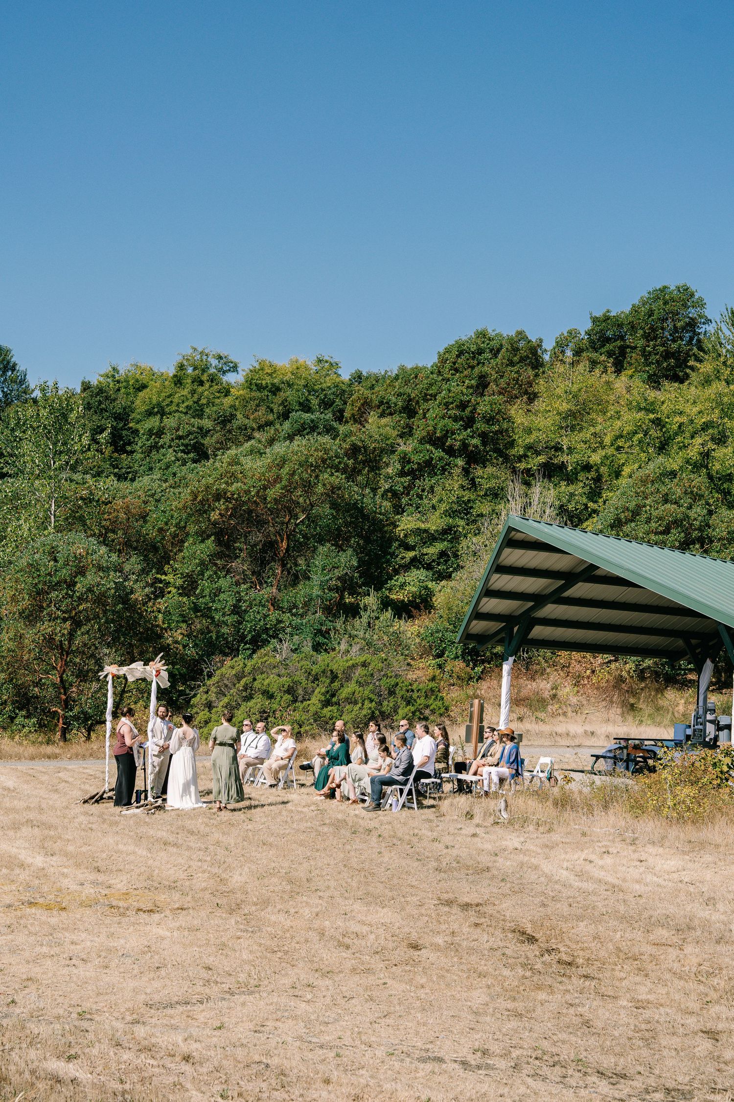Outdoor wedding ceremony setup with chairs and canopies on a grassy hillside under bright blue summer sky.