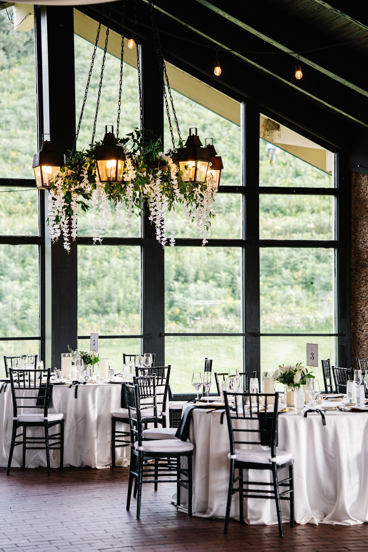 Elegant wedding reception venue with floor-to-ceiling windows, white tablecloths, black chiavari chairs and hanging chandeliers.