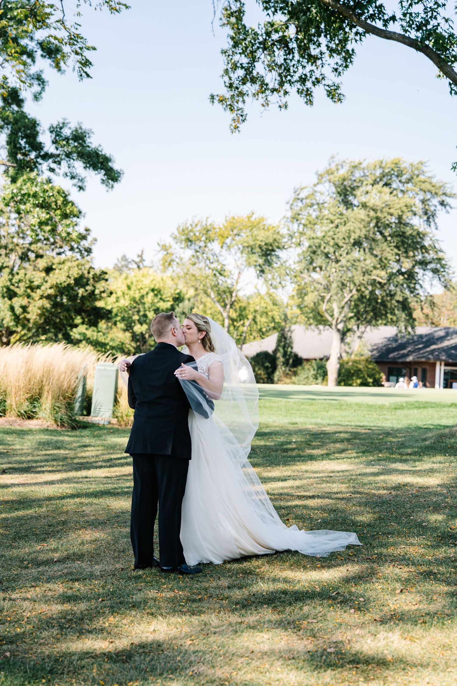 A couple shares an intimate moment on their wedding day in a sun-dappled garden setting with lush trees overhead.