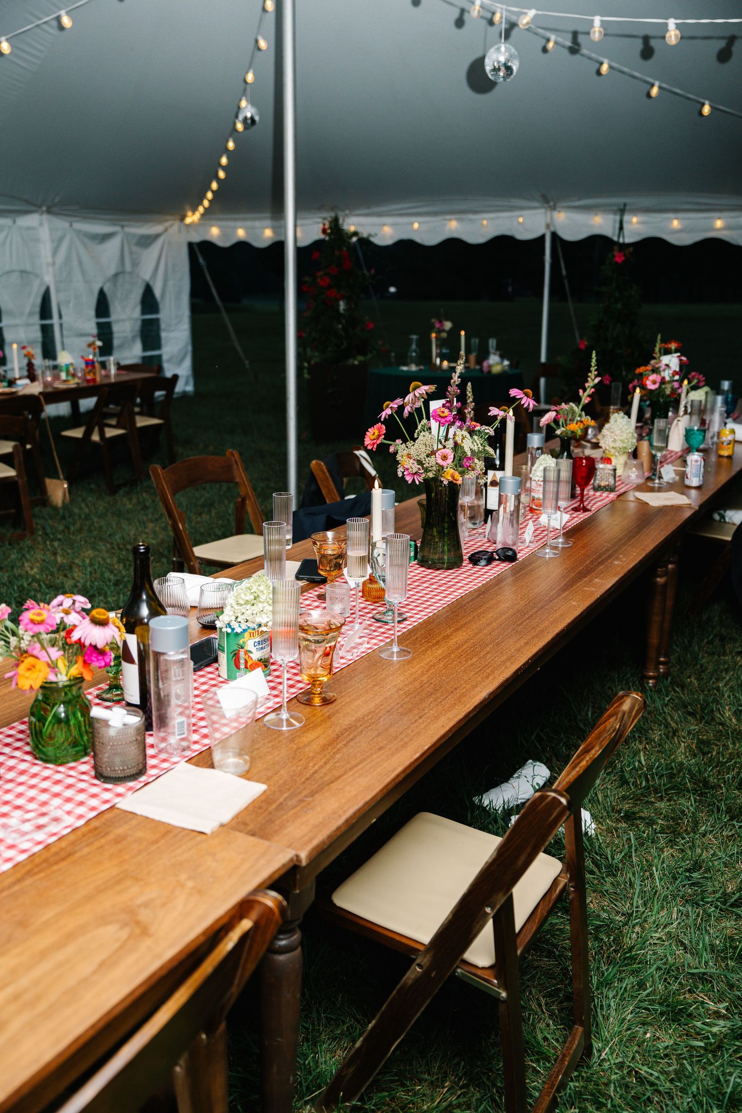 Long wooden banquet tables with red checkered tablecloths and string lights set up outdoors under a white tent.