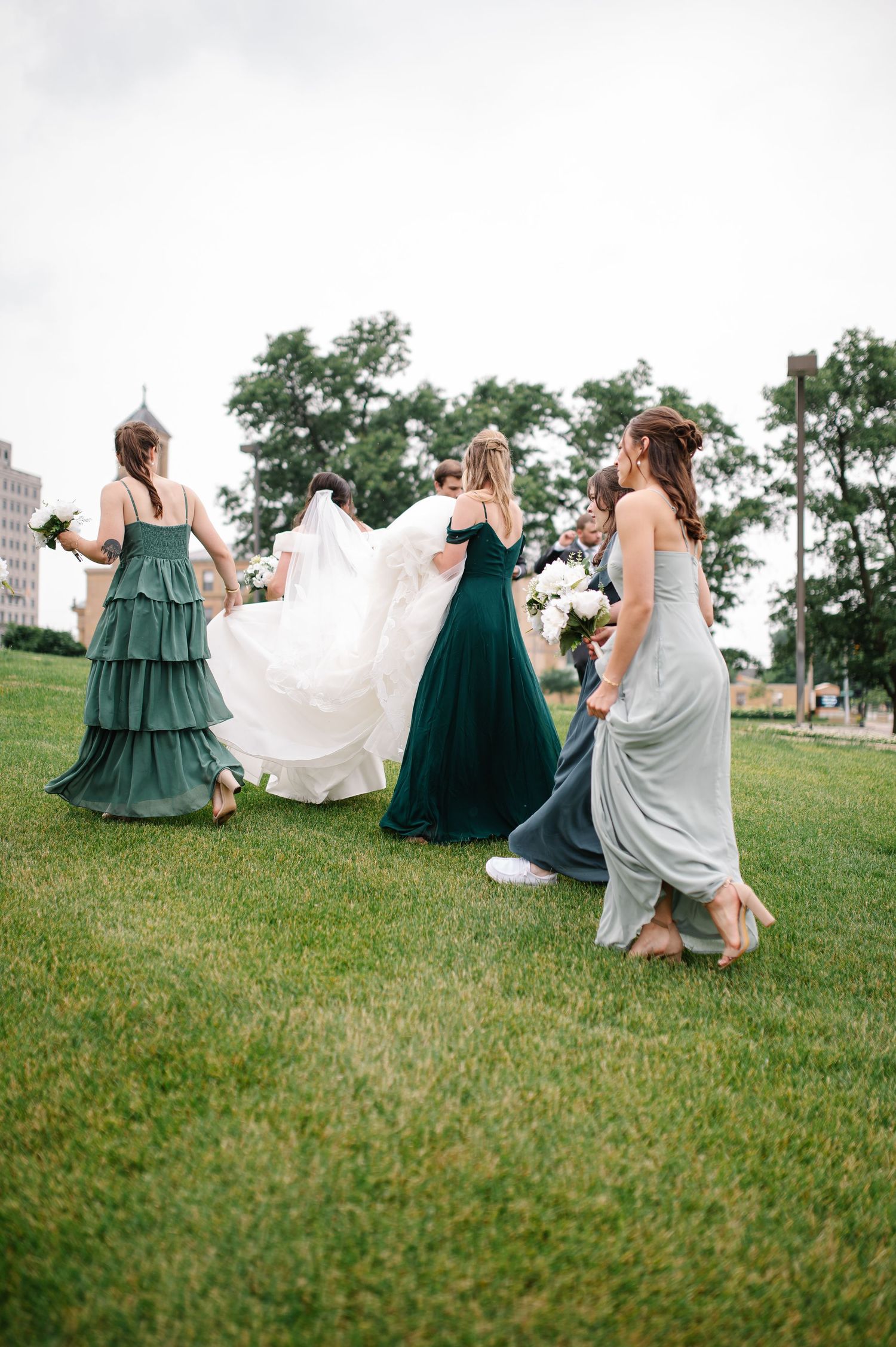 Bridesmaids in green and gray dresses walk across lawn holding wedding dress train on overcast day.