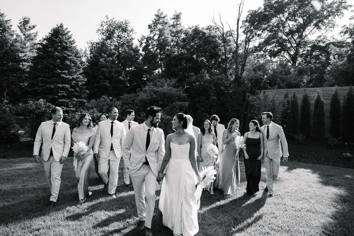 A wedding party in white and light colored formal attire walks together outdoors on a sunny day.