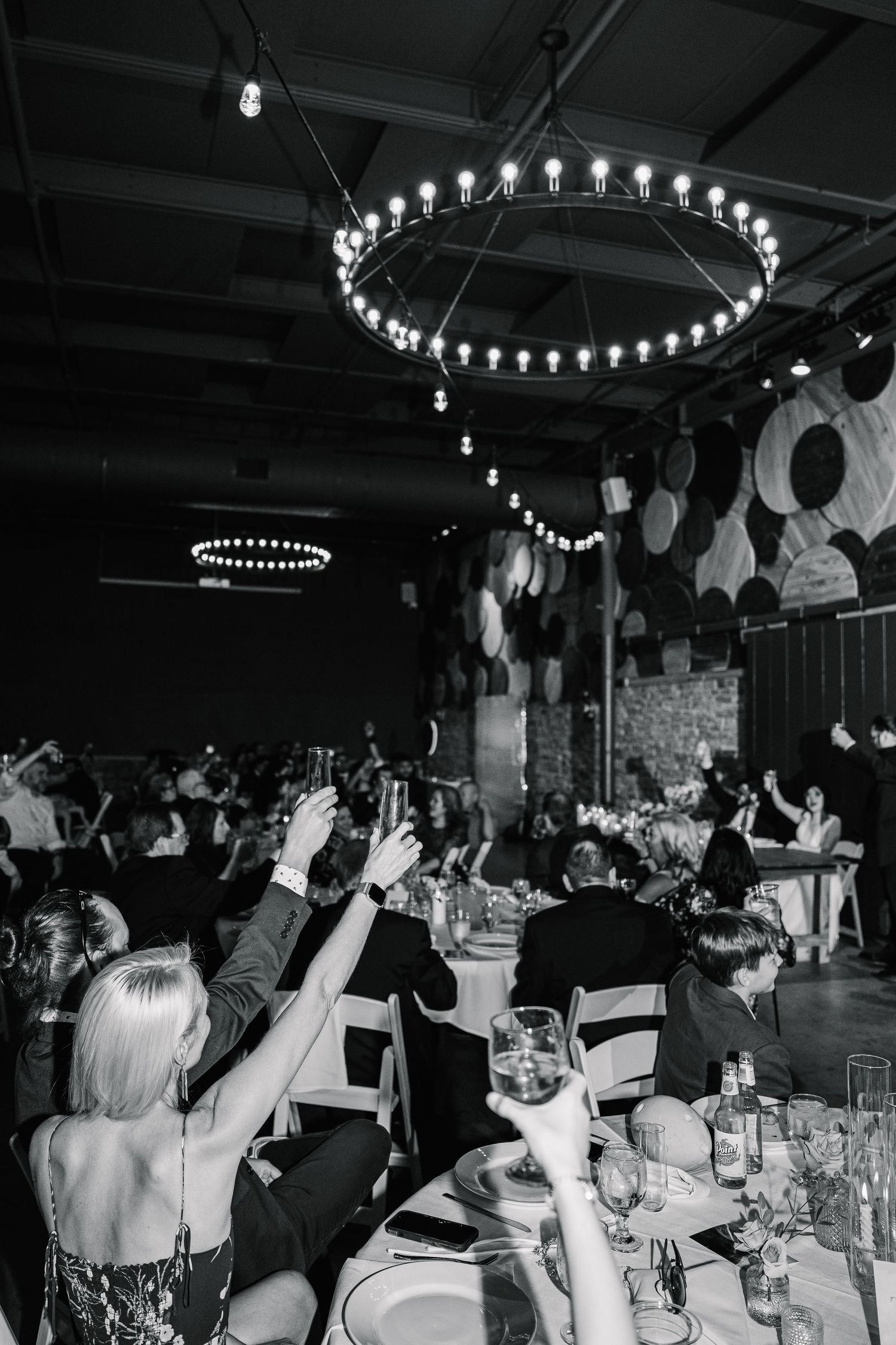 Wedding reception guests celebrate with raised arms under circular pendant lighting in a dimly lit venue.