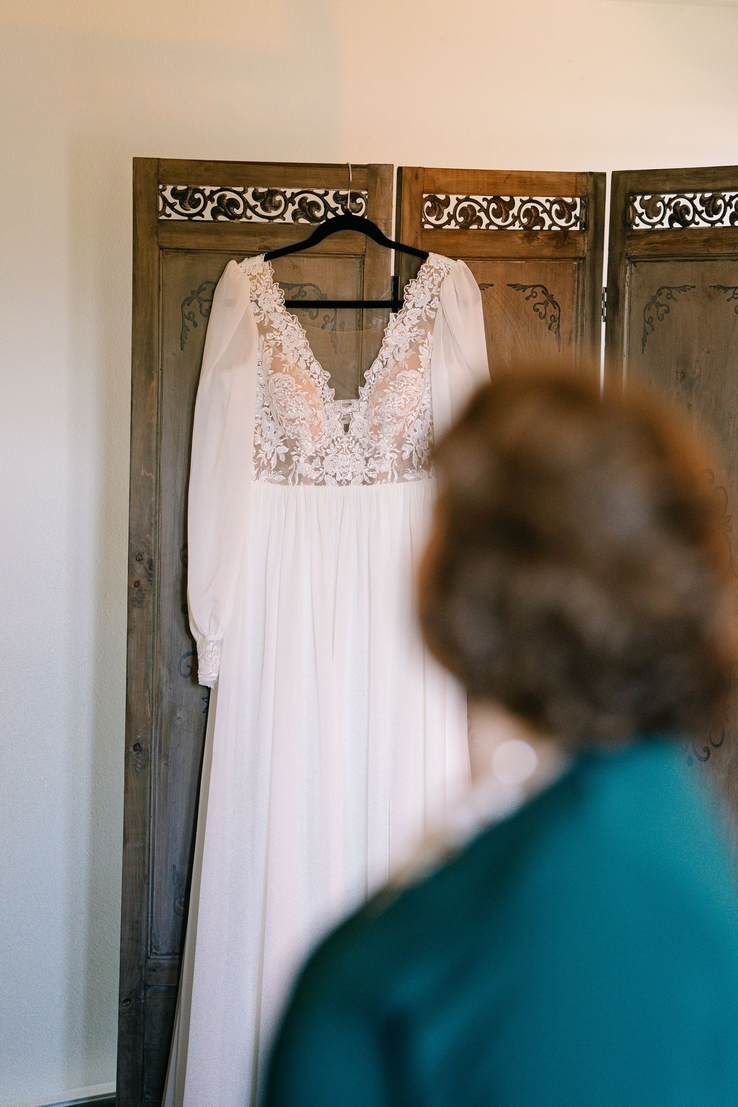 A white lace wedding dress hangs elegantly on a decorative room divider screen against a light colored wall.