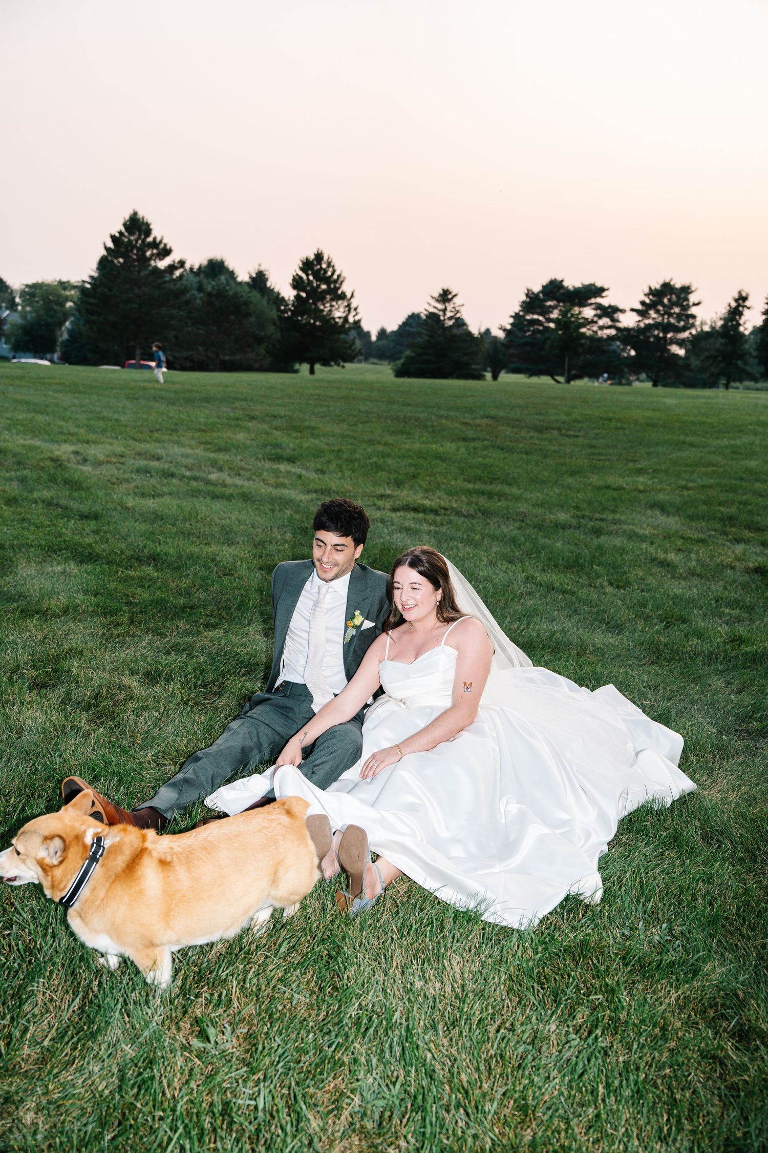 A couple in wedding attire sits on grass with their Corgi dog during sunset at a golf course.