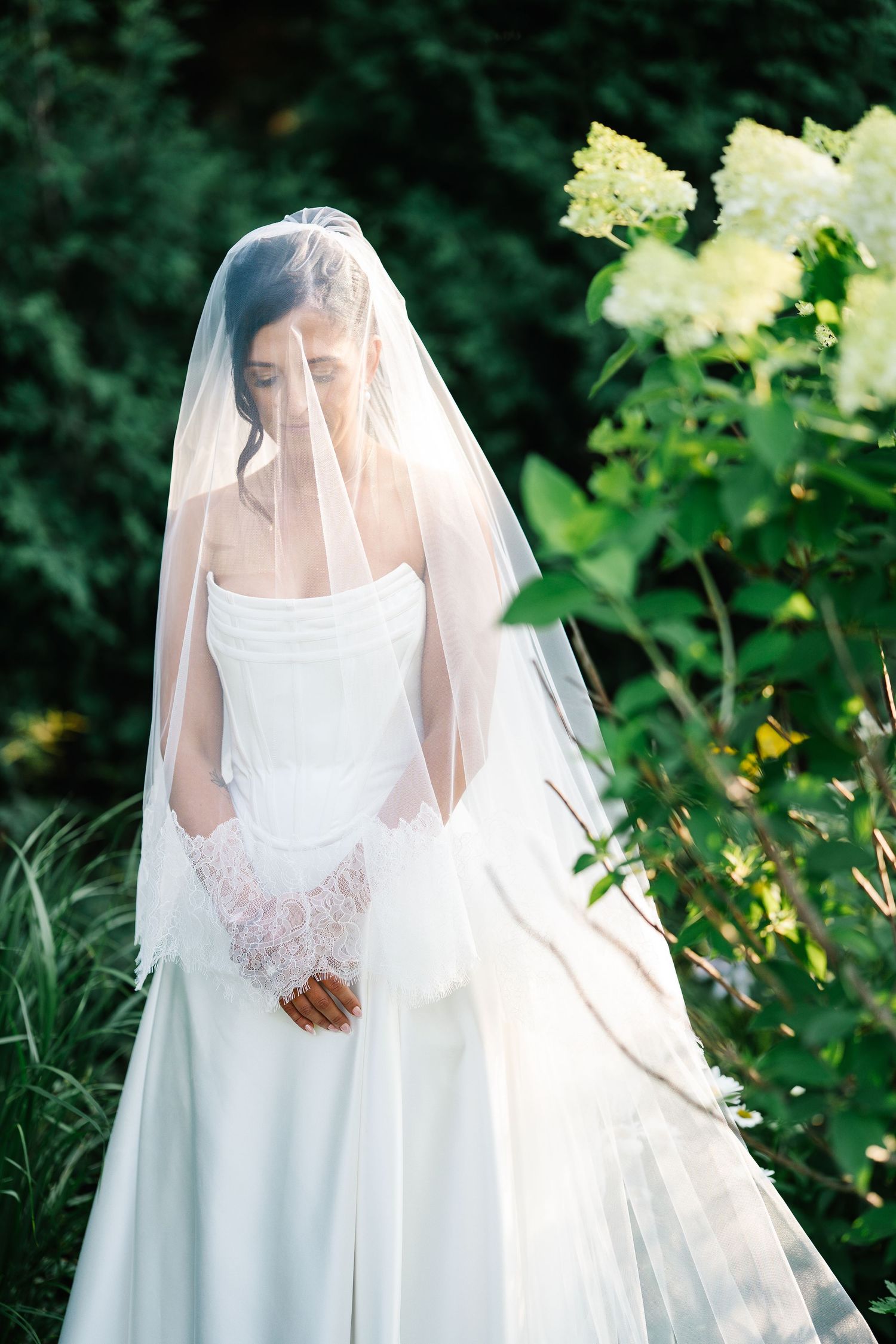 A bride in a flowing white wedding dress and veil stands in a lush garden setting with natural greenery.