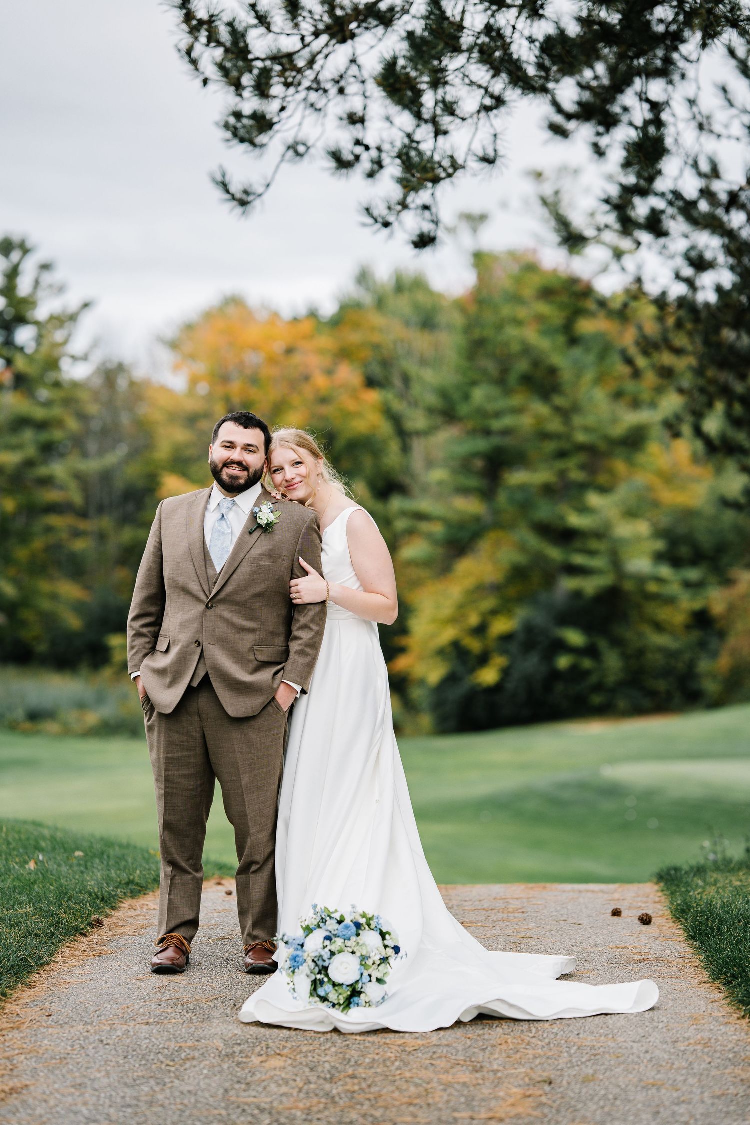 A wedding couple poses together on a gravel path with autumn trees and green grass in the background.