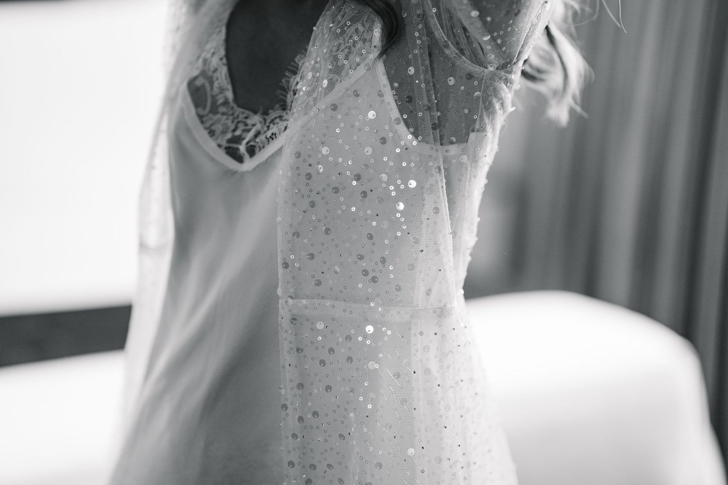 A delicate bridal gown with sparkling beaded details and sheer mesh overlay hangs elegantly against a light background.