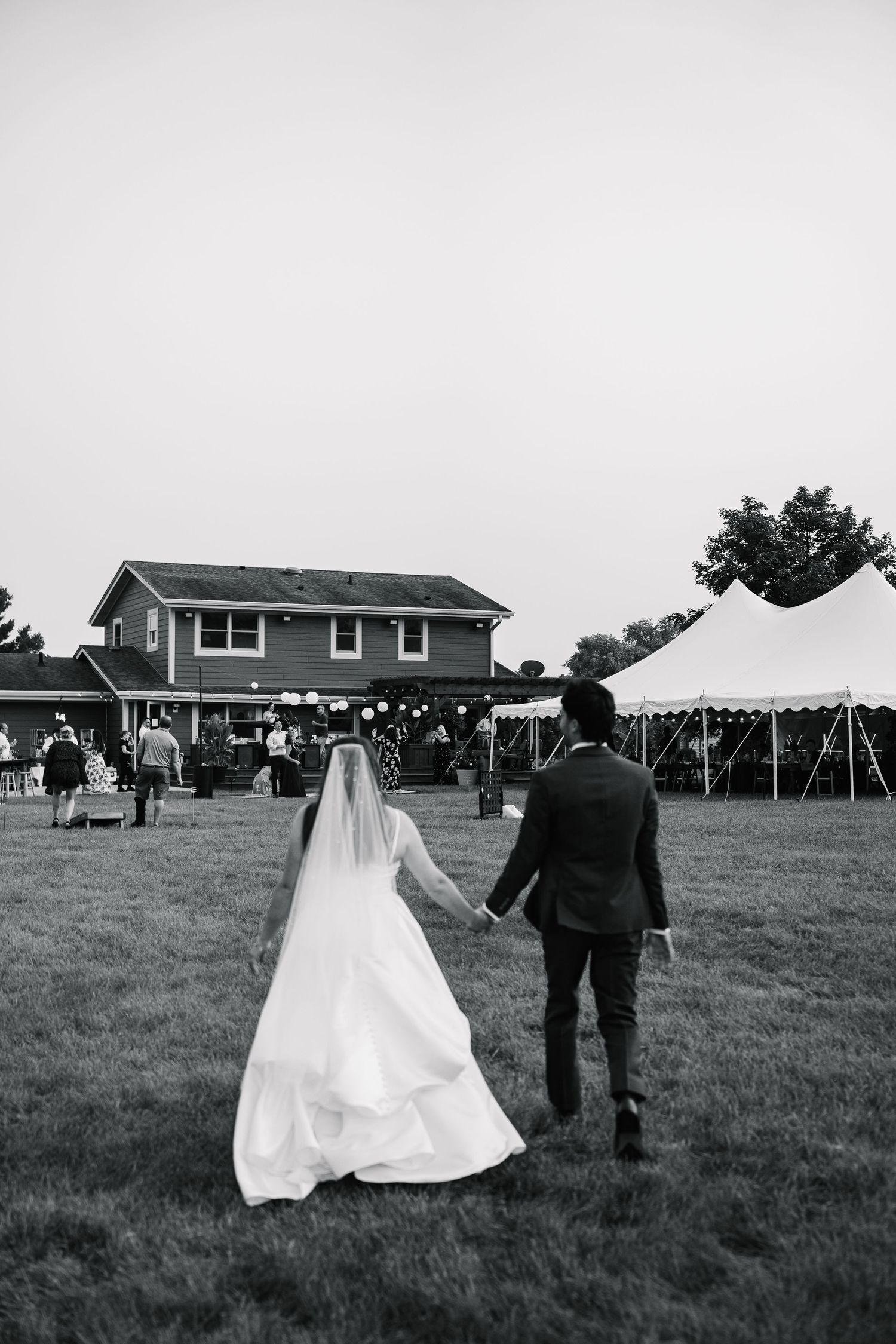 Black and white wedding photo sequence showing a couple walking together across a grassy field with tents and buildings in background.
