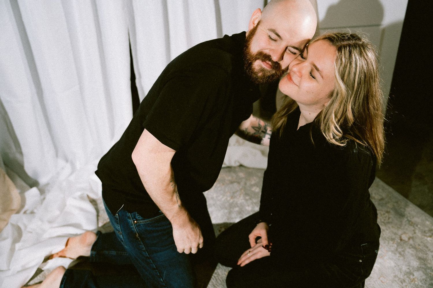 A couple in matching black tops share a tender moment while sitting together indoors.