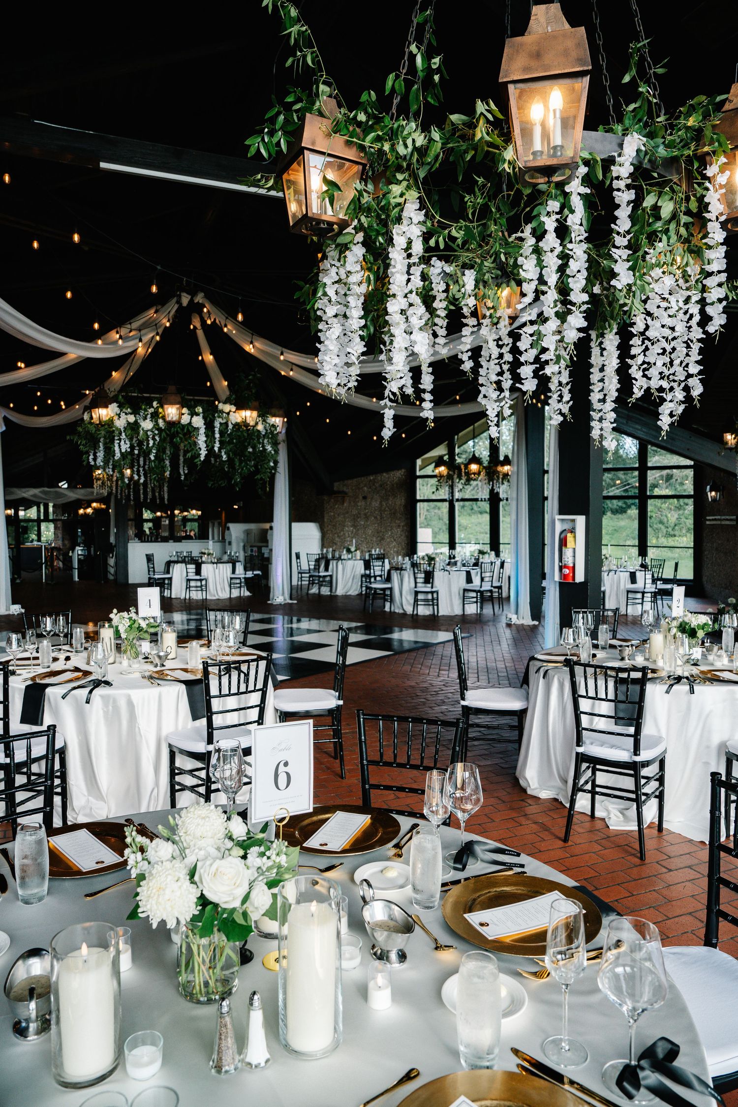 Elegant wedding reception venue with hanging white wisteria, copper lanterns, and round tables set with white linens and candles.