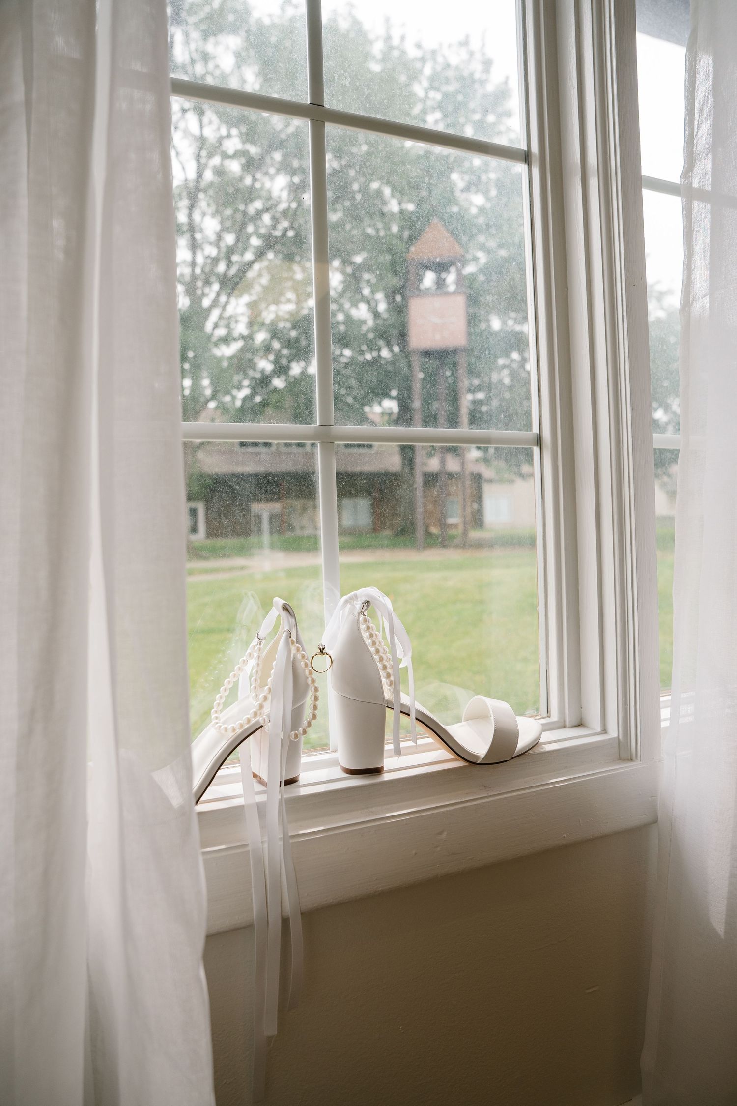 White bridal shoes rest on a windowsill with a birdhouse visible through the window pane in the background.