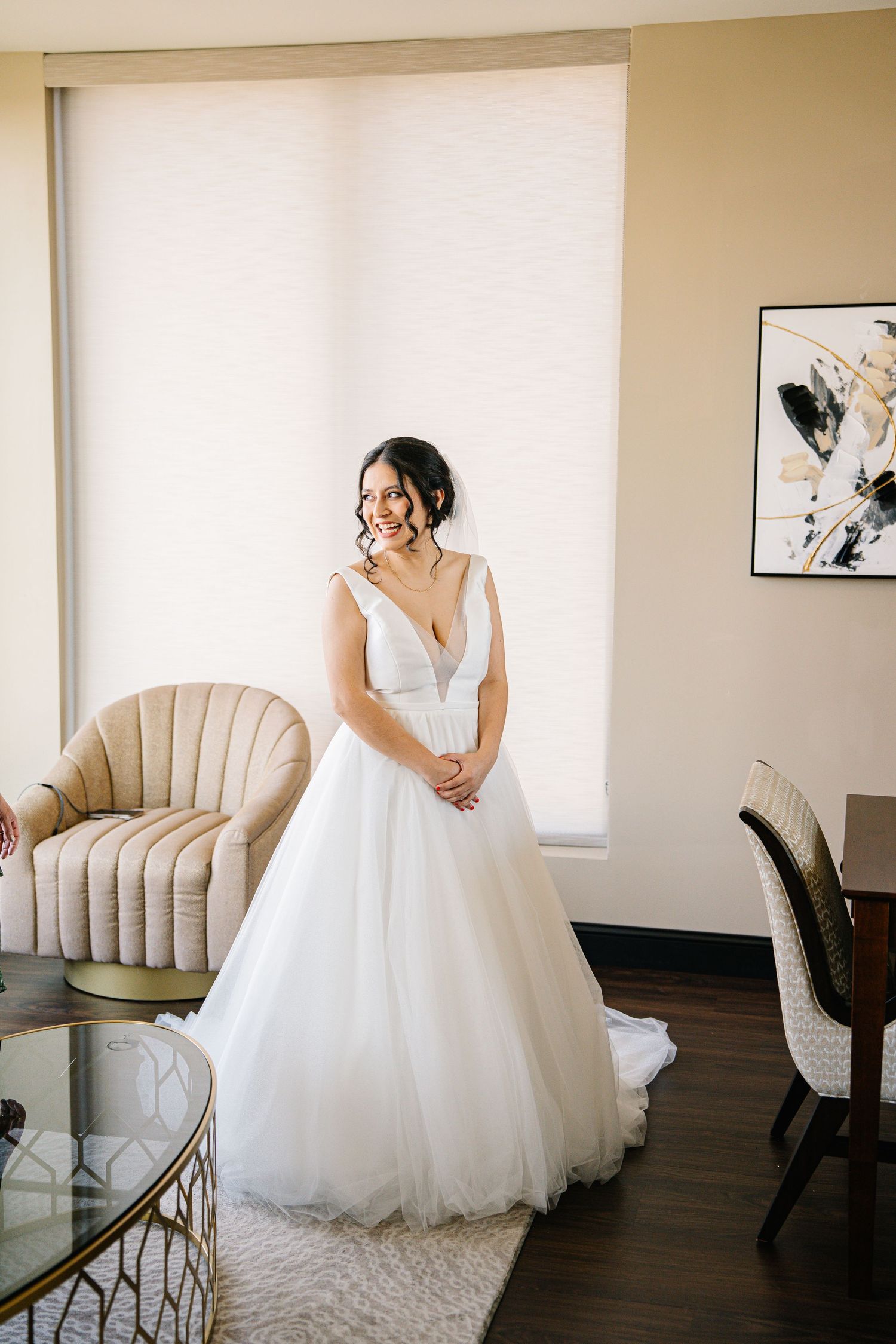 A flowing white wedding gown is showcased in an elegant room with beige walls and plush furniture.