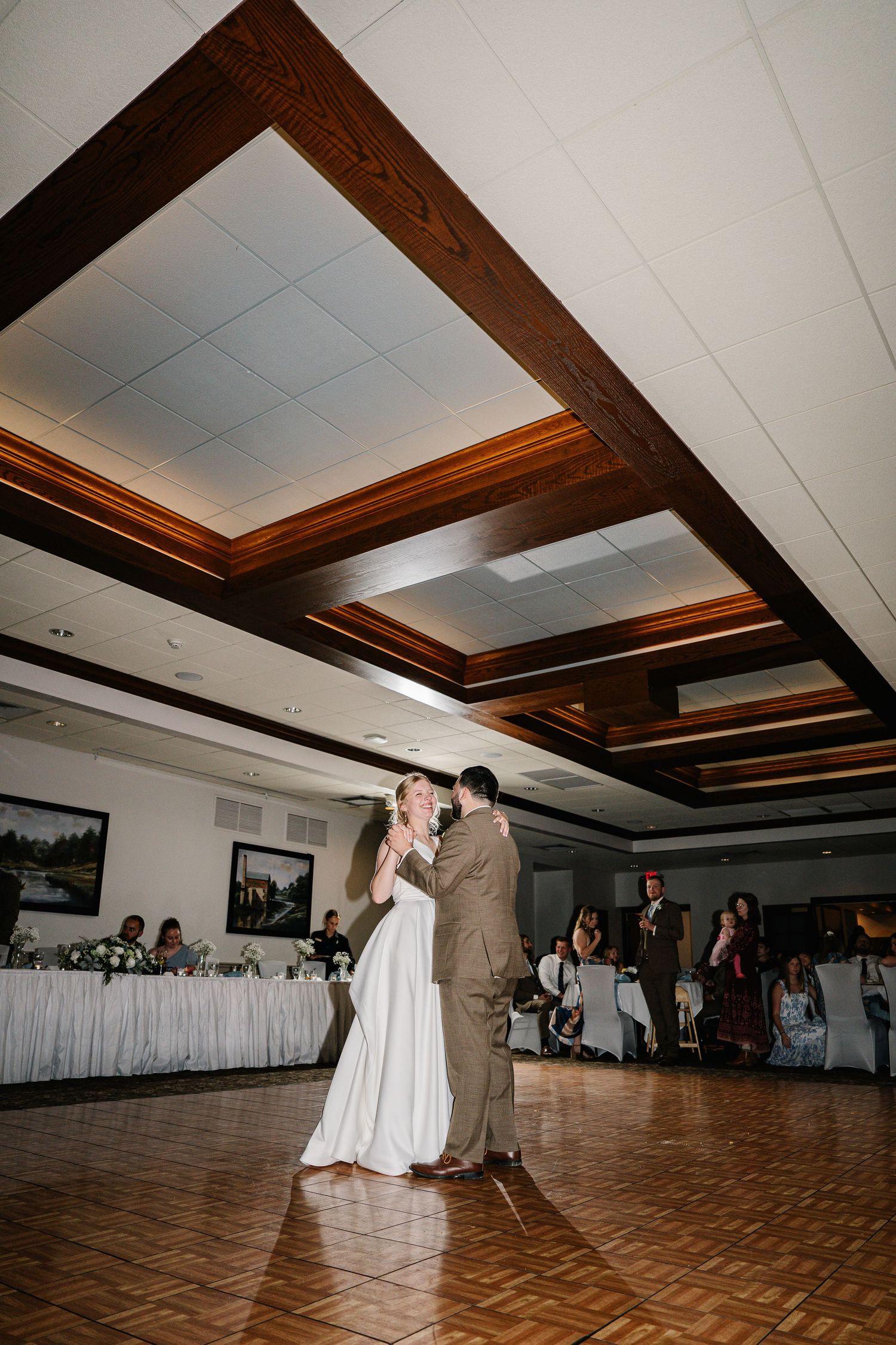 A couple shares their first dance on a parquet floor in a banquet hall with wooden ceiling beams.