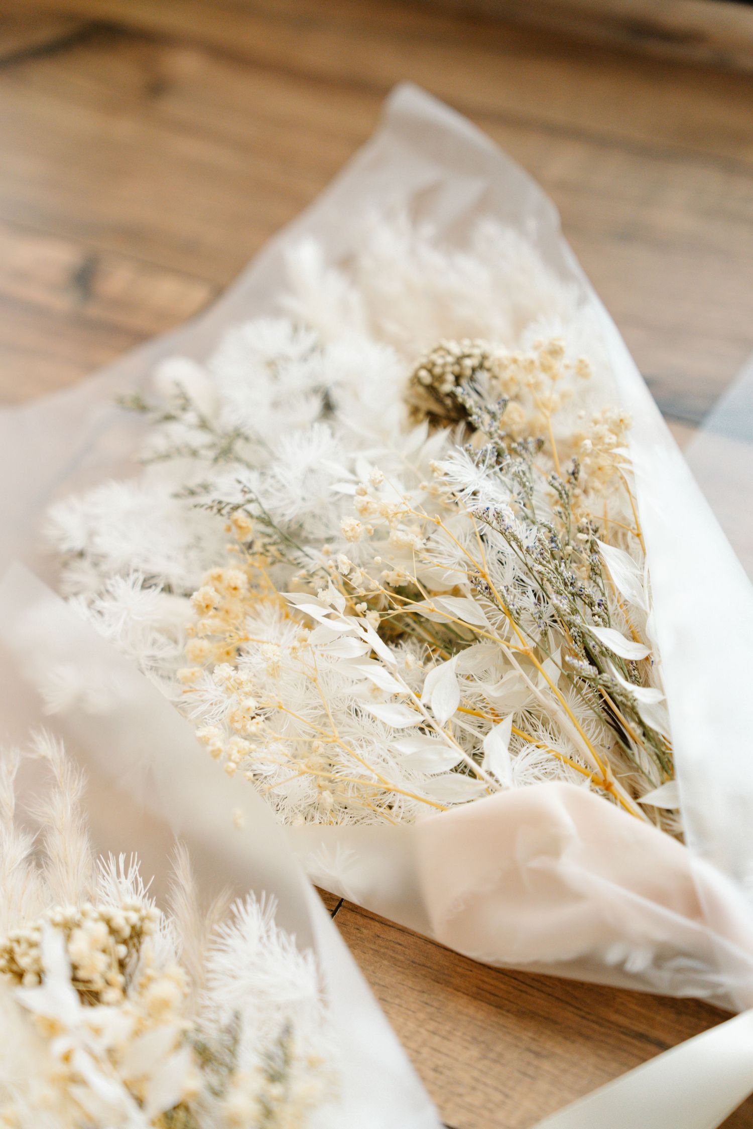 Delicate dried flowers and grasses wrapped in white paper create a soft, rustic arrangement on wooden surface.