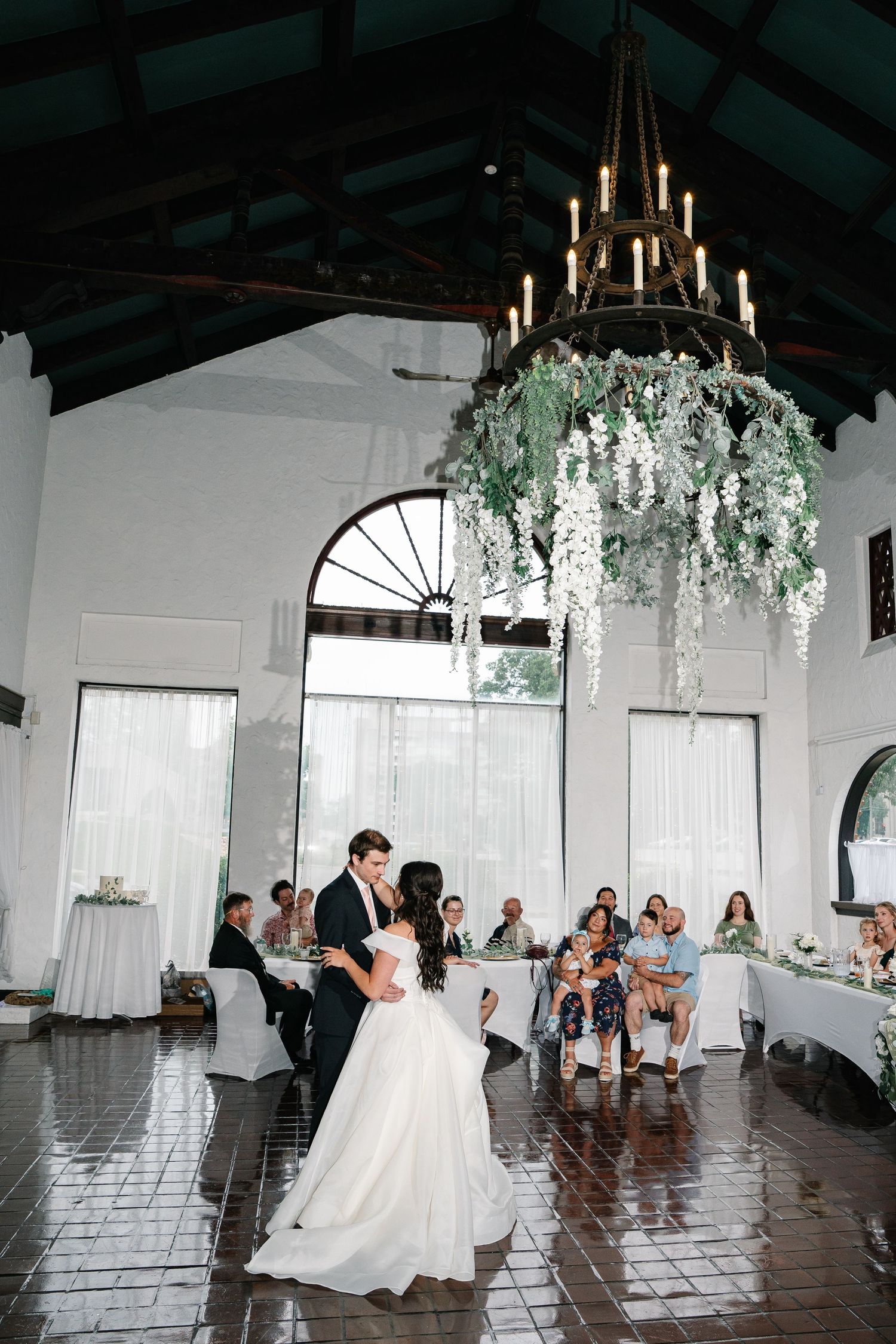 Couple shares their first dance in an elegant white reception venue decorated with hanging floral arrangements and chandeliers.