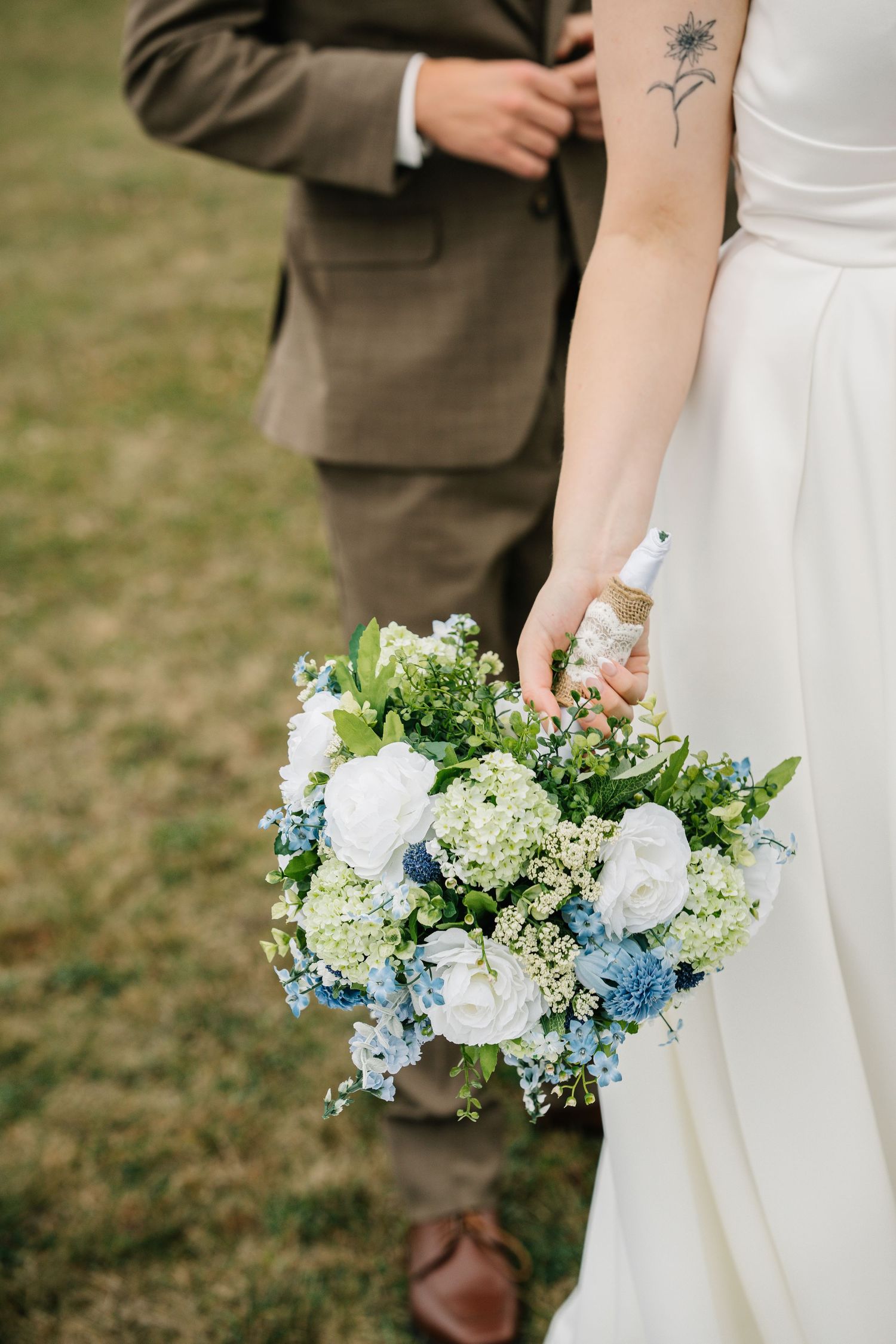 A delicate wedding bouquet featuring white roses, blue hydrangeas, and greenery held against a white dress.