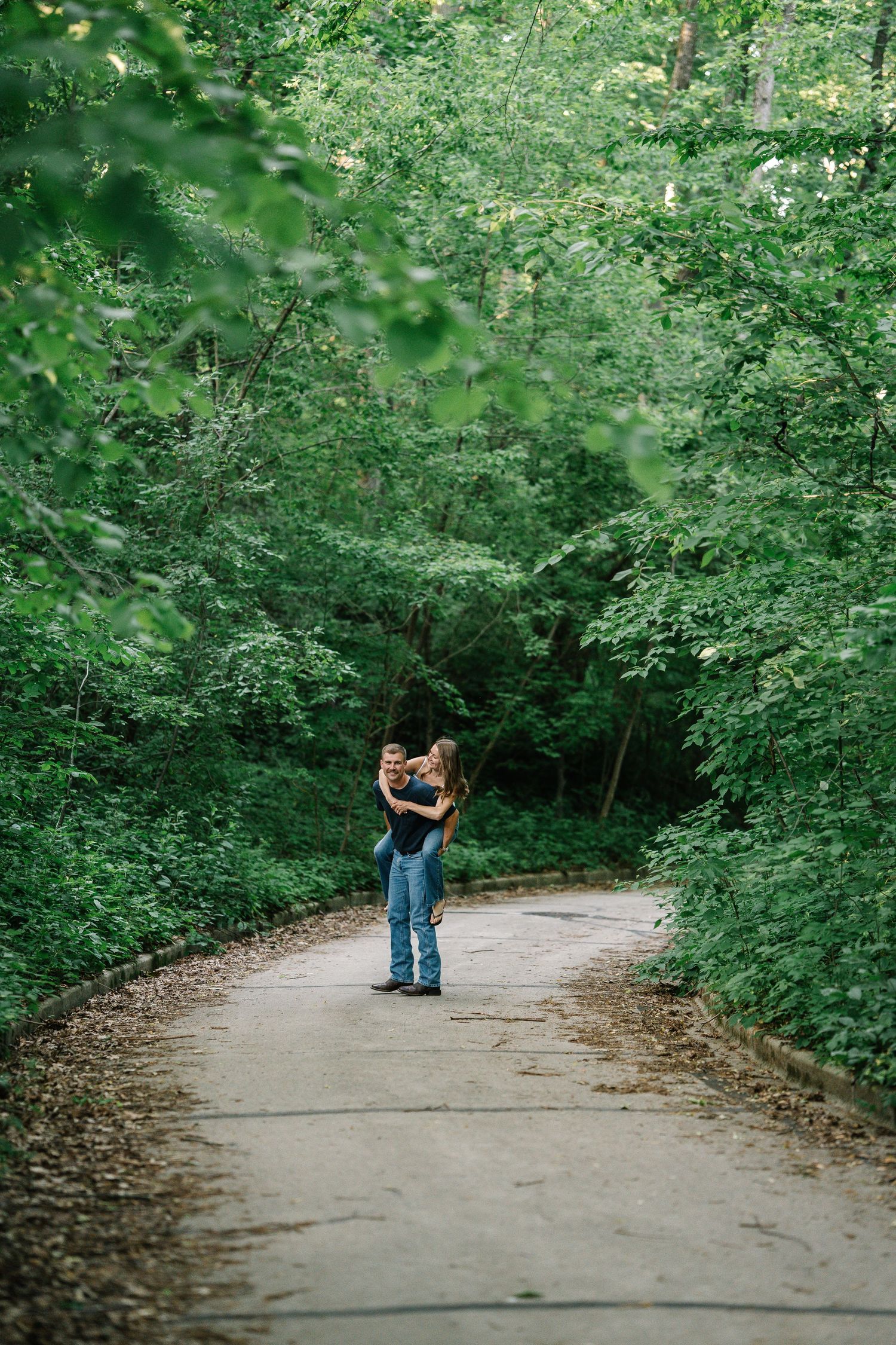 Couple embracing on a winding forest path surrounded by lush green trees in a serene outdoor setting.