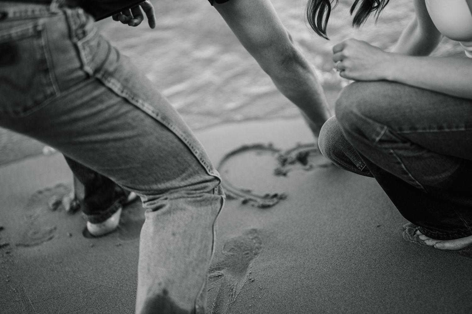 Black and white photo of couple drawing a heart in beach sand, wearing jeans, romantic seaside moment.