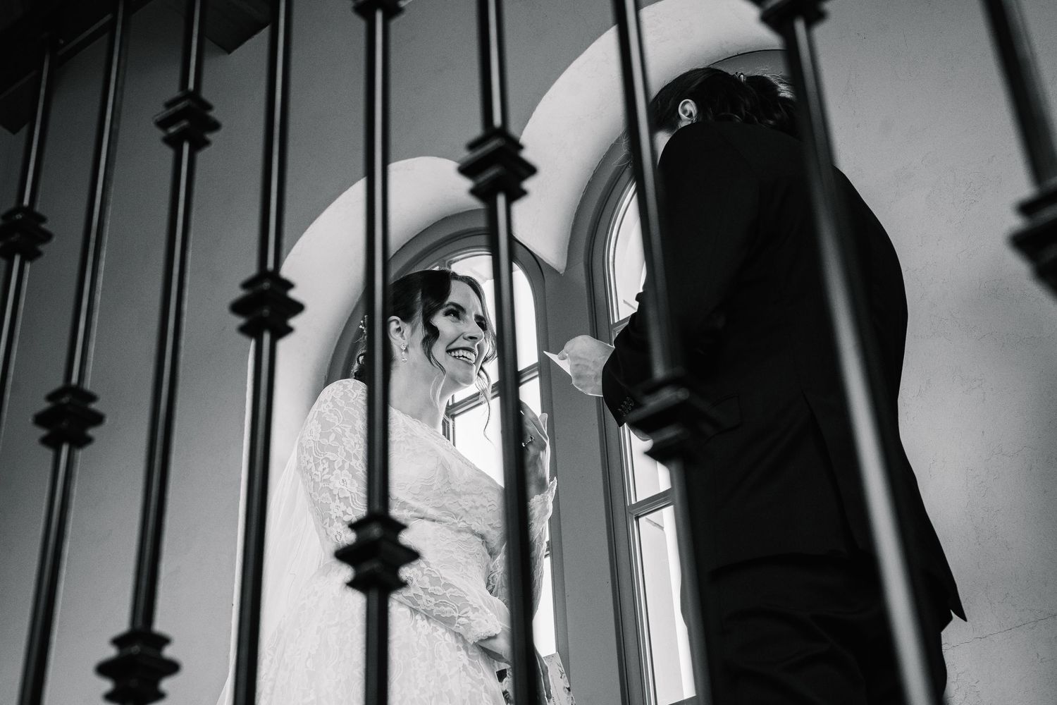 Black and white photo of a smiling bride in a wedding dress seen through ornate iron railings with groom.