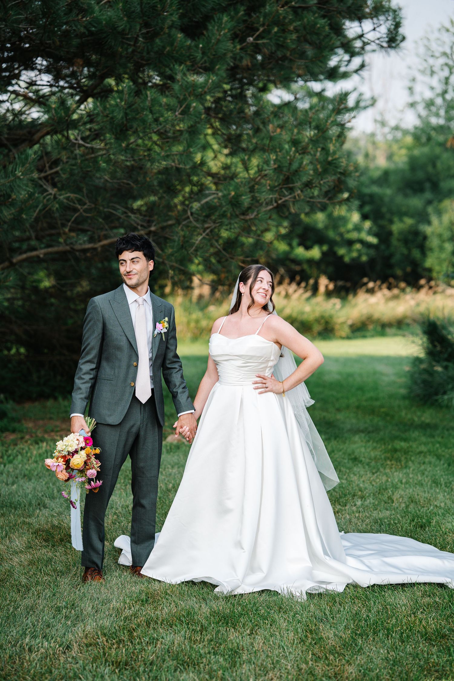 A couple in wedding attire poses together on a grassy lawn with evergreen trees in the background.