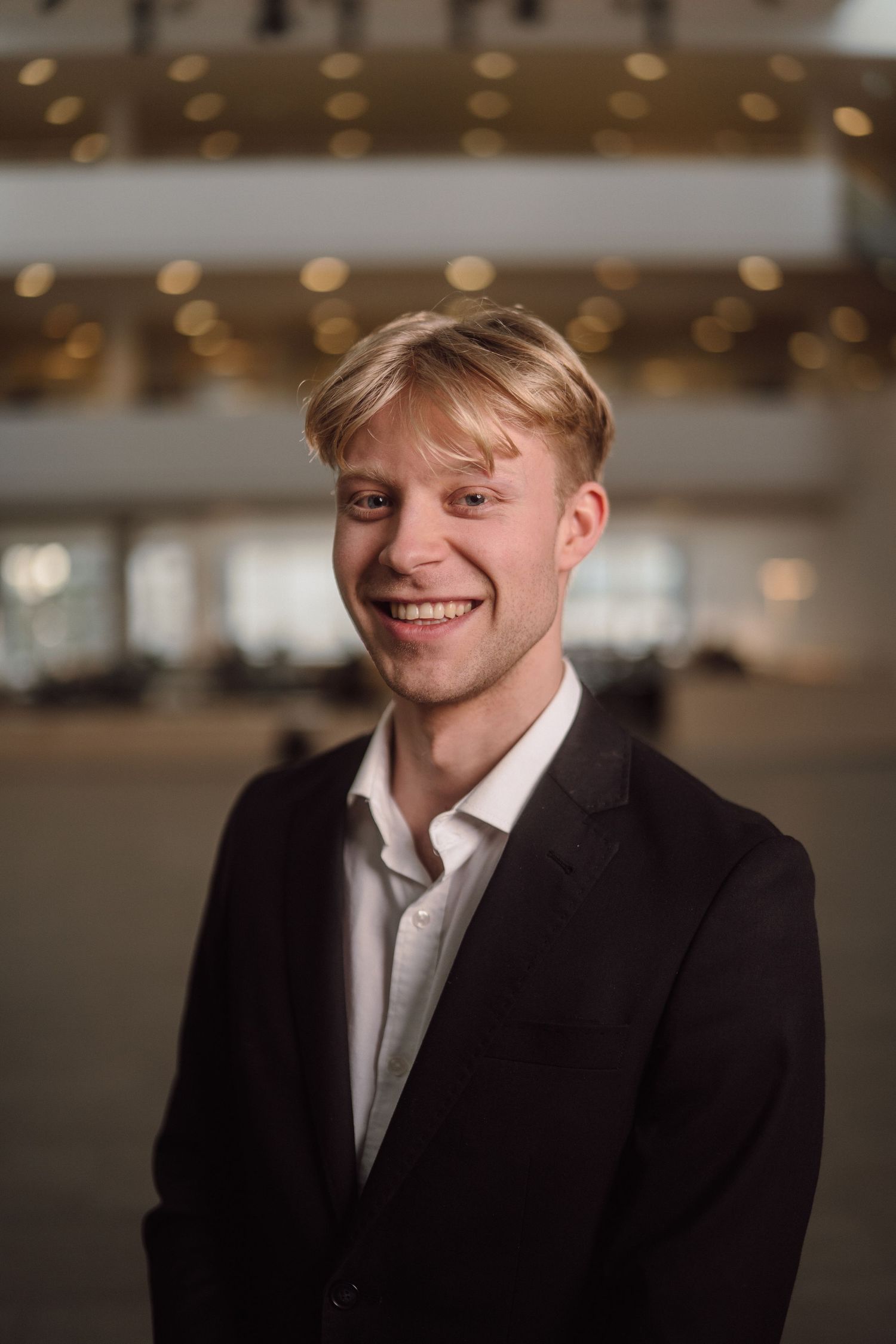 A professional headshot of someone in a dark suit and white shirt against a blurred interior background.