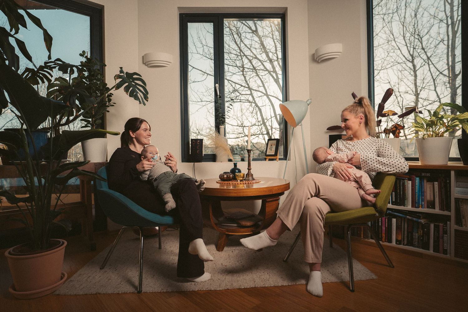Two mothers sit in cozy chairs holding infants in a plant-filled living room with large windows.