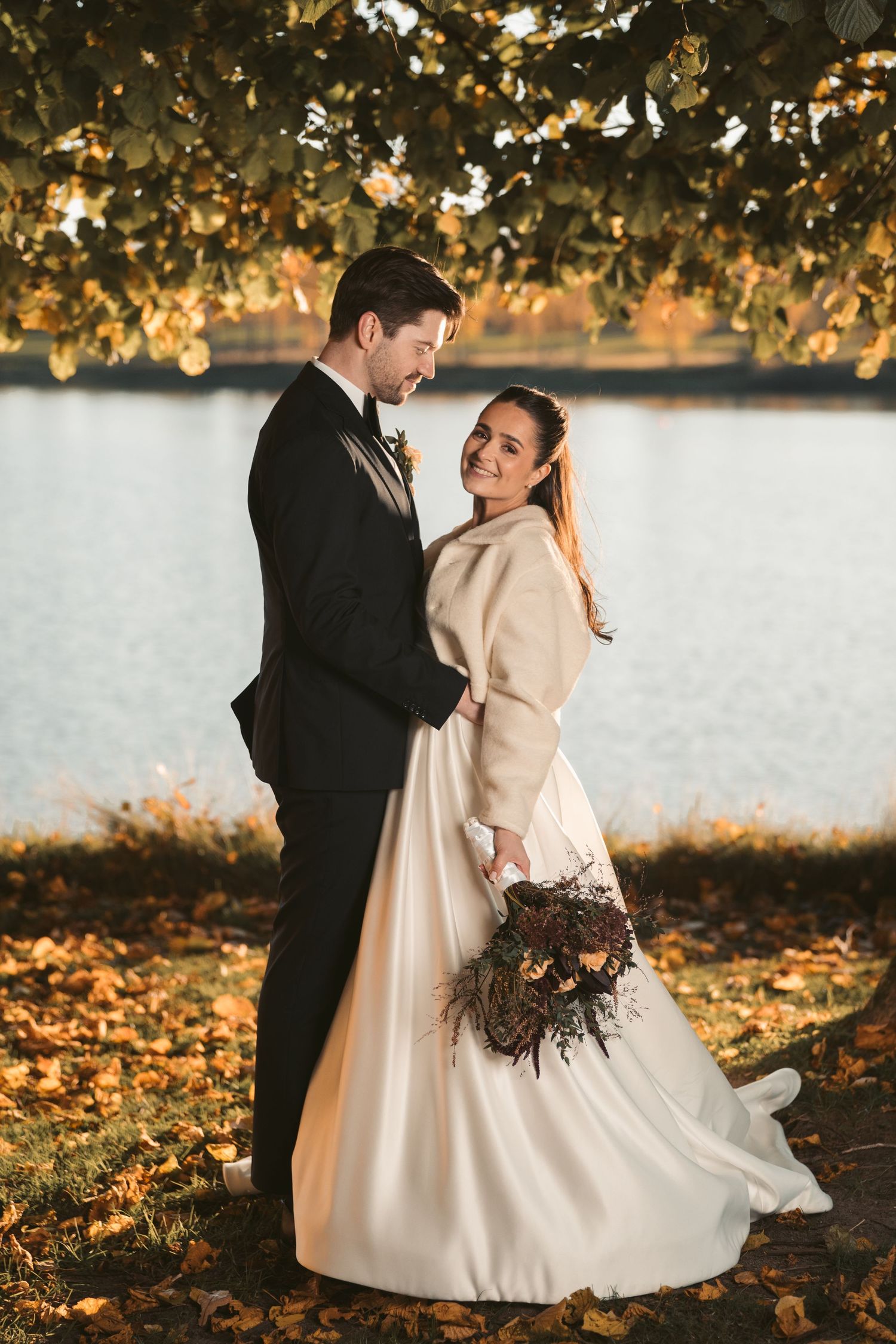 Romantic autumn wedding photoshoot by the lake with golden leaves and warm sunlight.