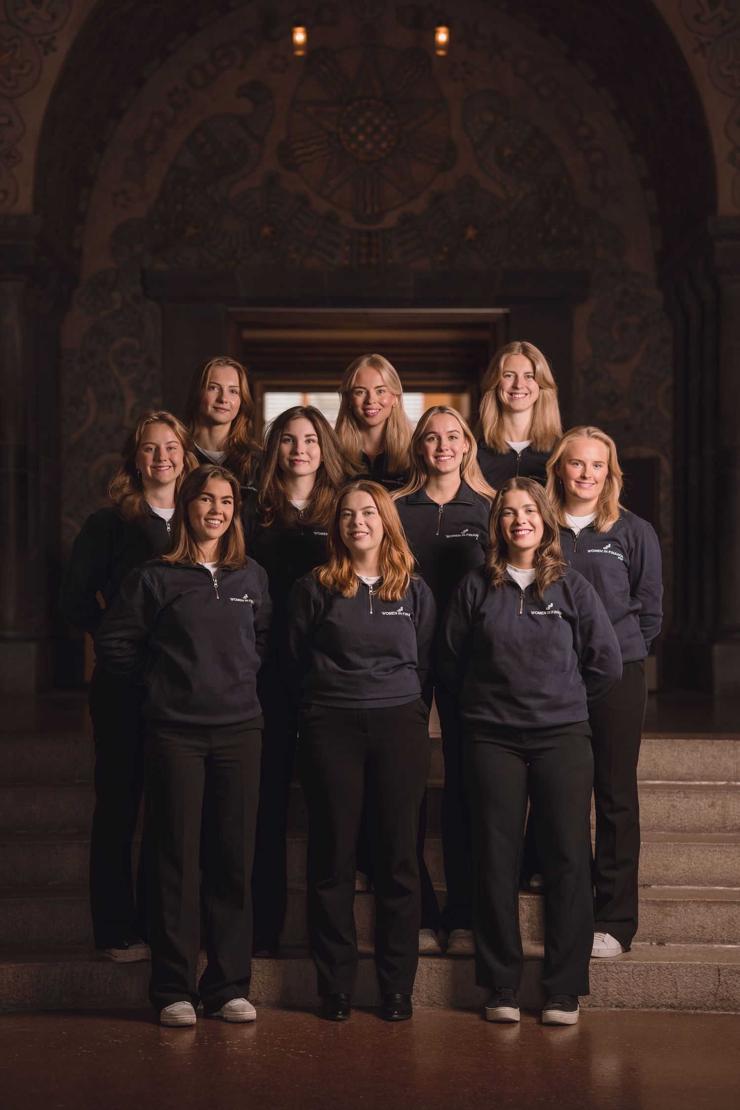 A group wearing matching dark uniforms poses together in an ornate stone archway with dim lighting.