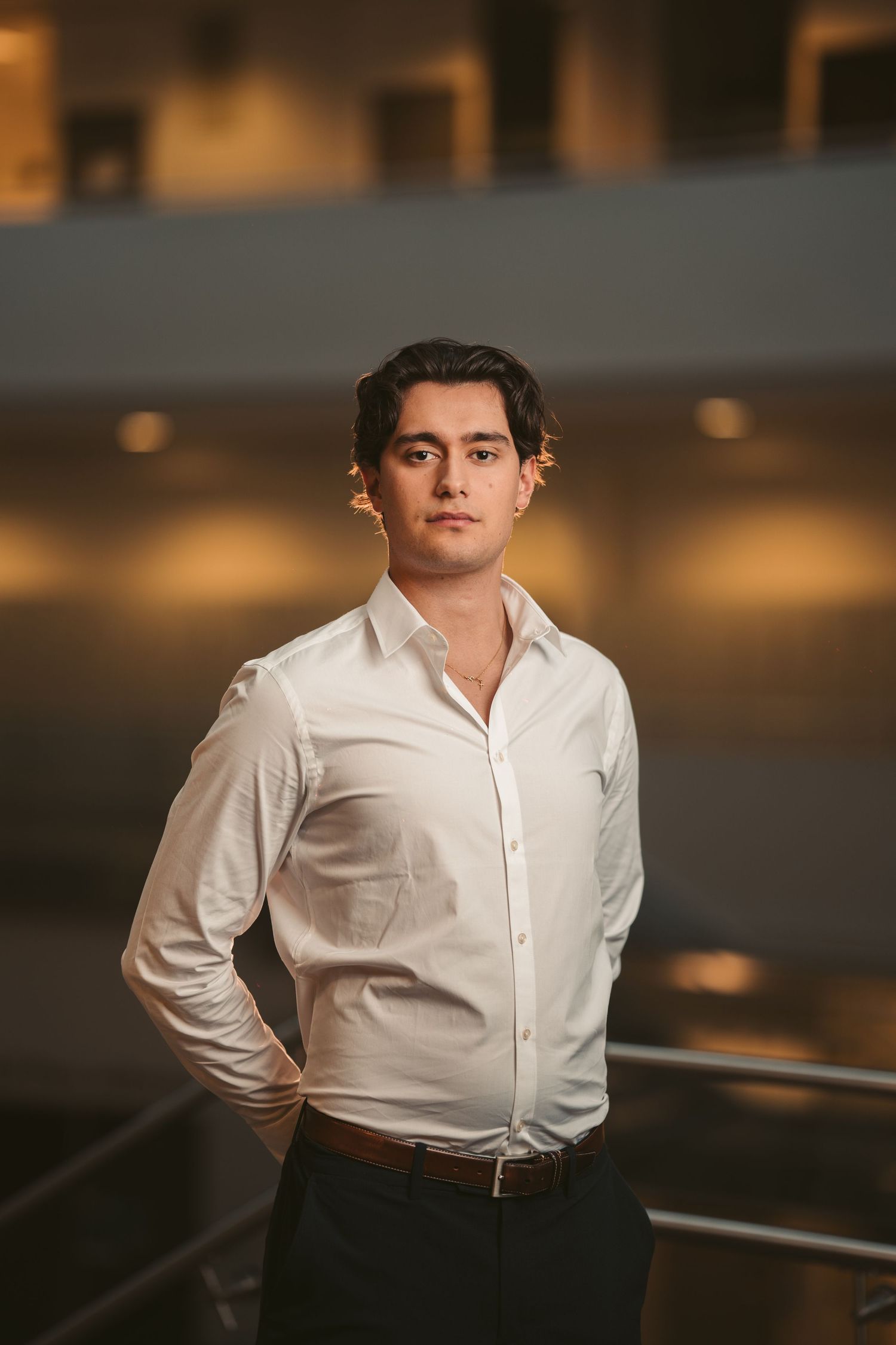 A person in a crisp white dress shirt and dark pants poses in a warmly lit indoor setting.
