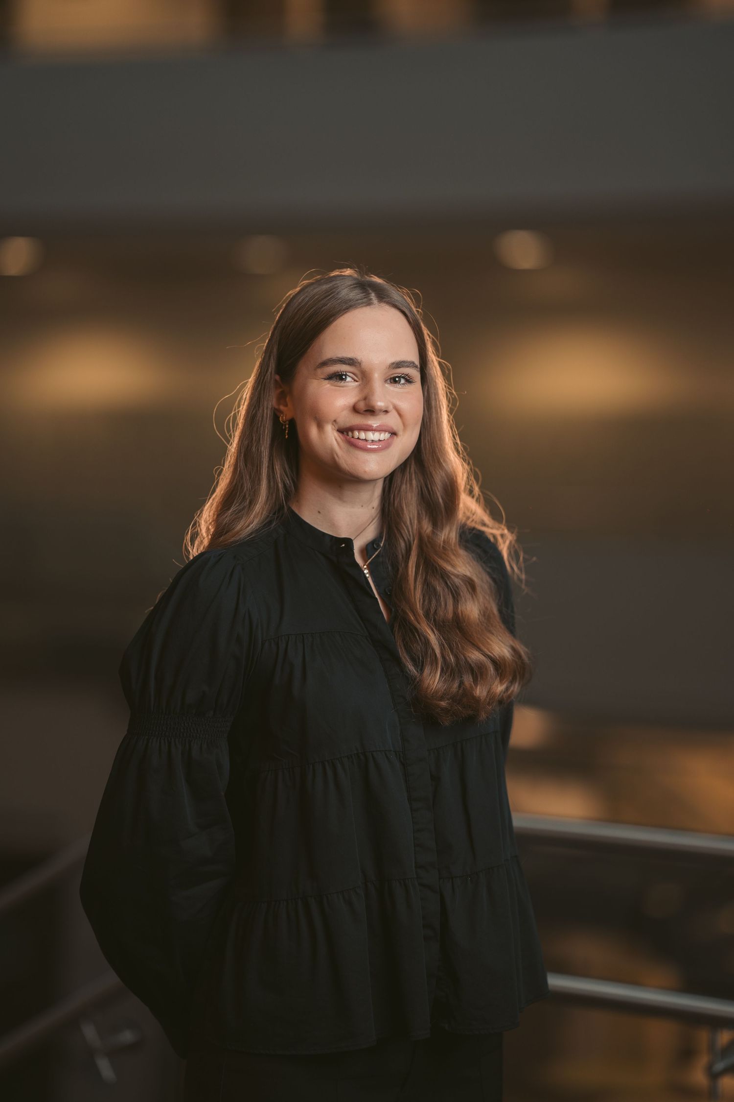 A person wearing a flowing black garment poses in front of a softly lit background.
