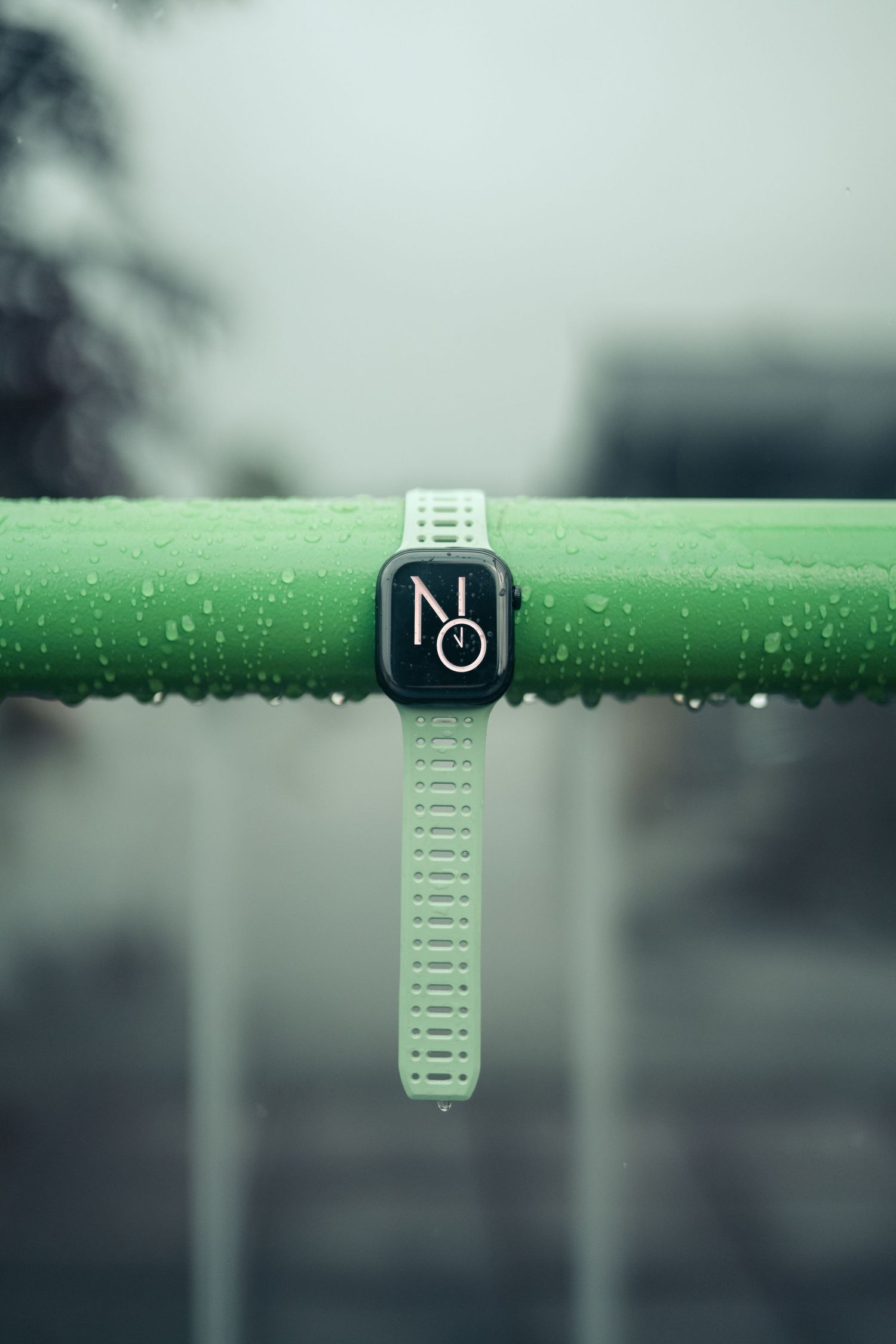 Apple Watch with white sport band displayed on a wet green railing in the rain, showcasing its water resistance.